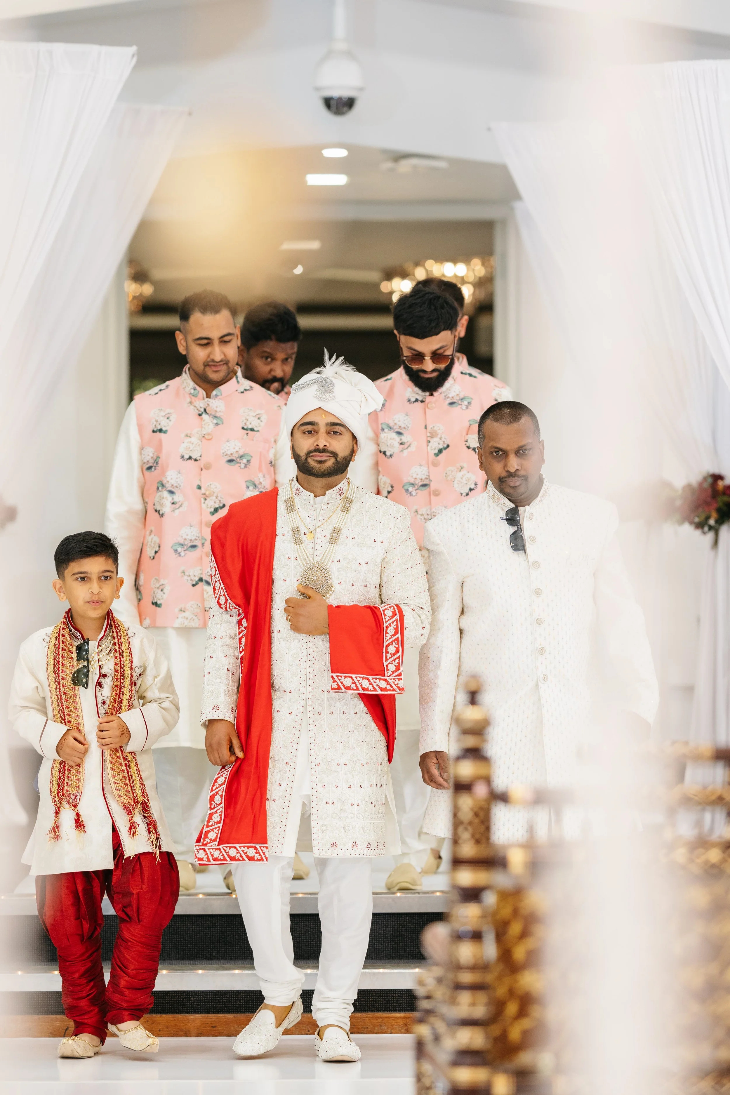 A group of Indian men and boys dressed in traditional attire, walking down a staircase at a celebration or wedding, with white curtains and chandeliers in the background.