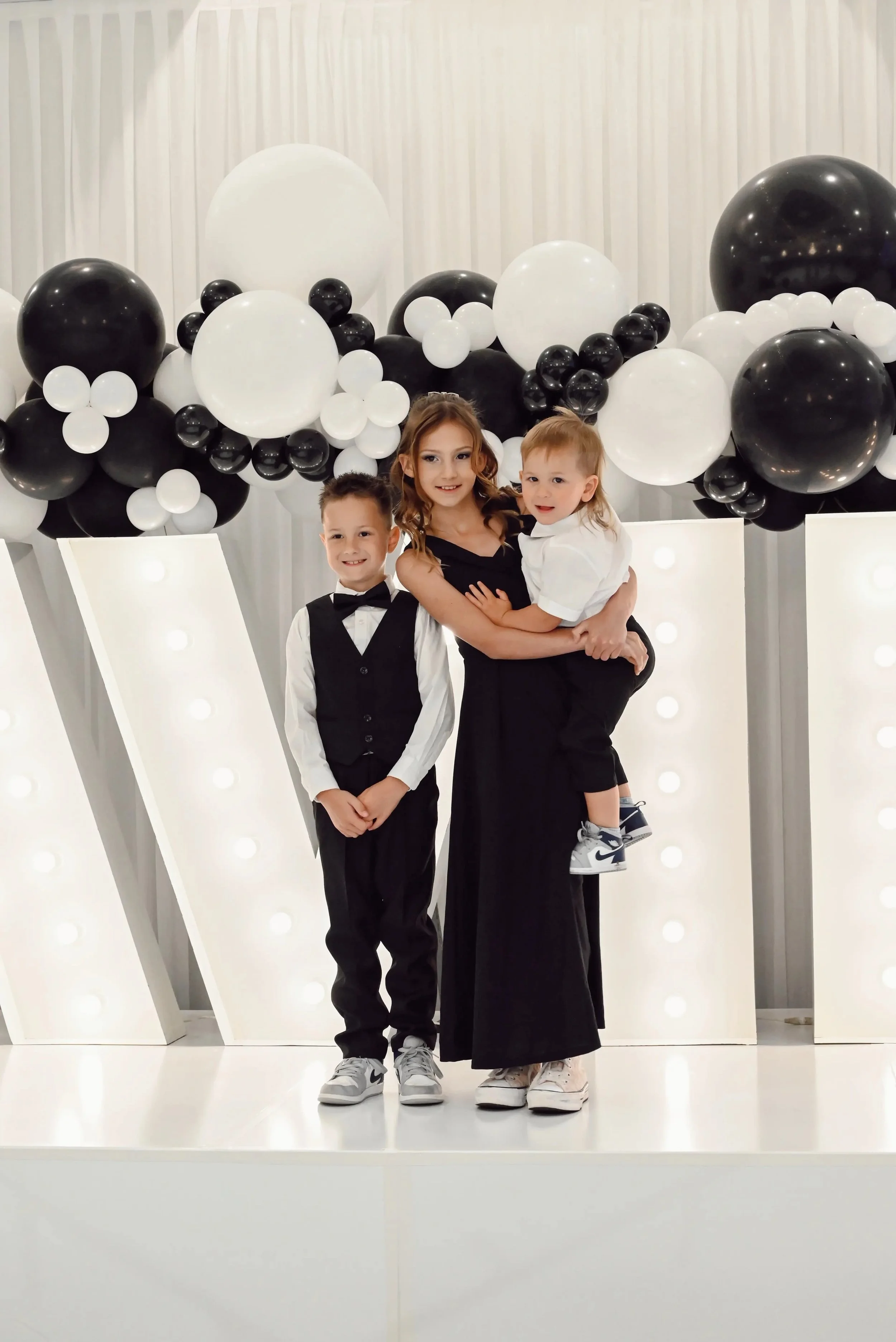 A woman holding a young girl, with a boy standing beside them, all dressed in formal attire, standing in front of black and white balloon decorations.