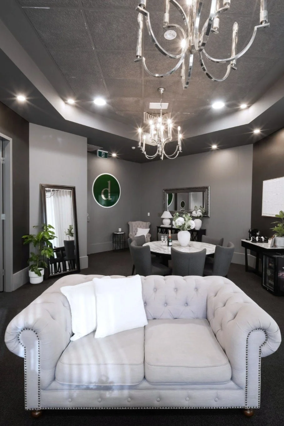 A cozy living room with a plush cream-colored tufted sofa in the foreground, a round dining table with gray chairs and white floral centerpiece in the background, a large mirror on the wall, and elegant chandeliers with multiple candle-like lights ha