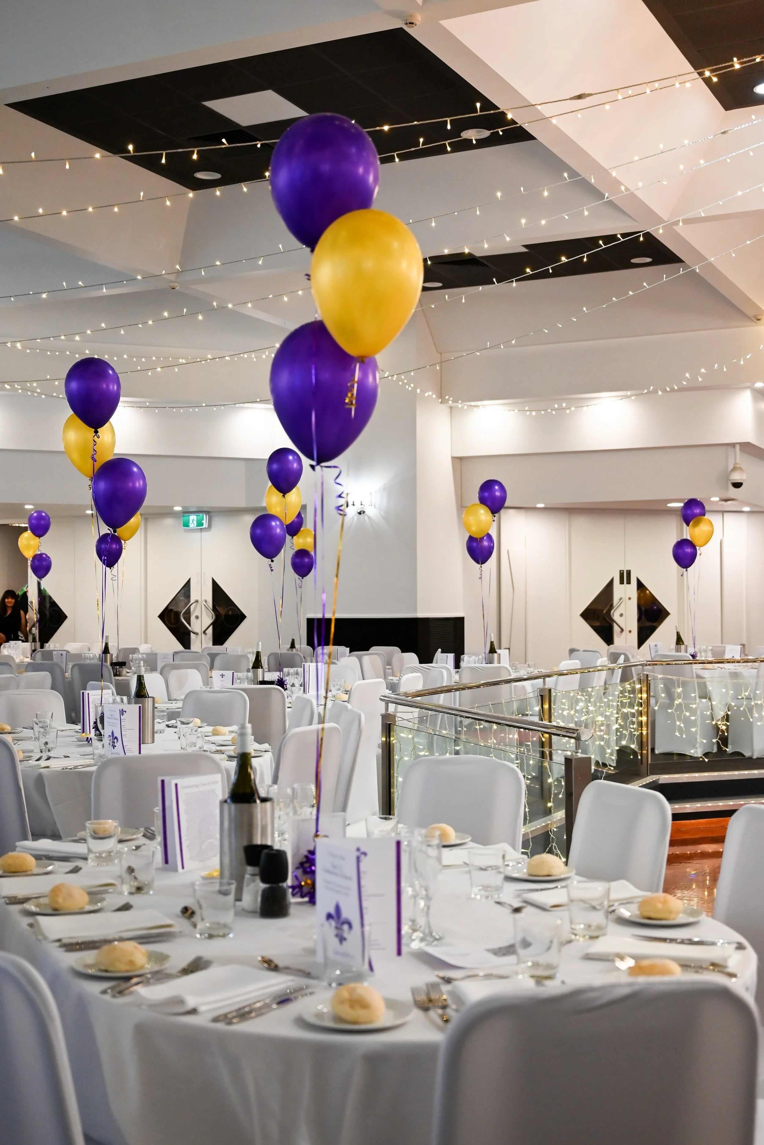 A decorated banquet hall with round tables covered in white tablecloths, set with place settings, bread rolls, and purple and gold balloons. The ceiling is decorated with string lights, creating a festive atmosphere.