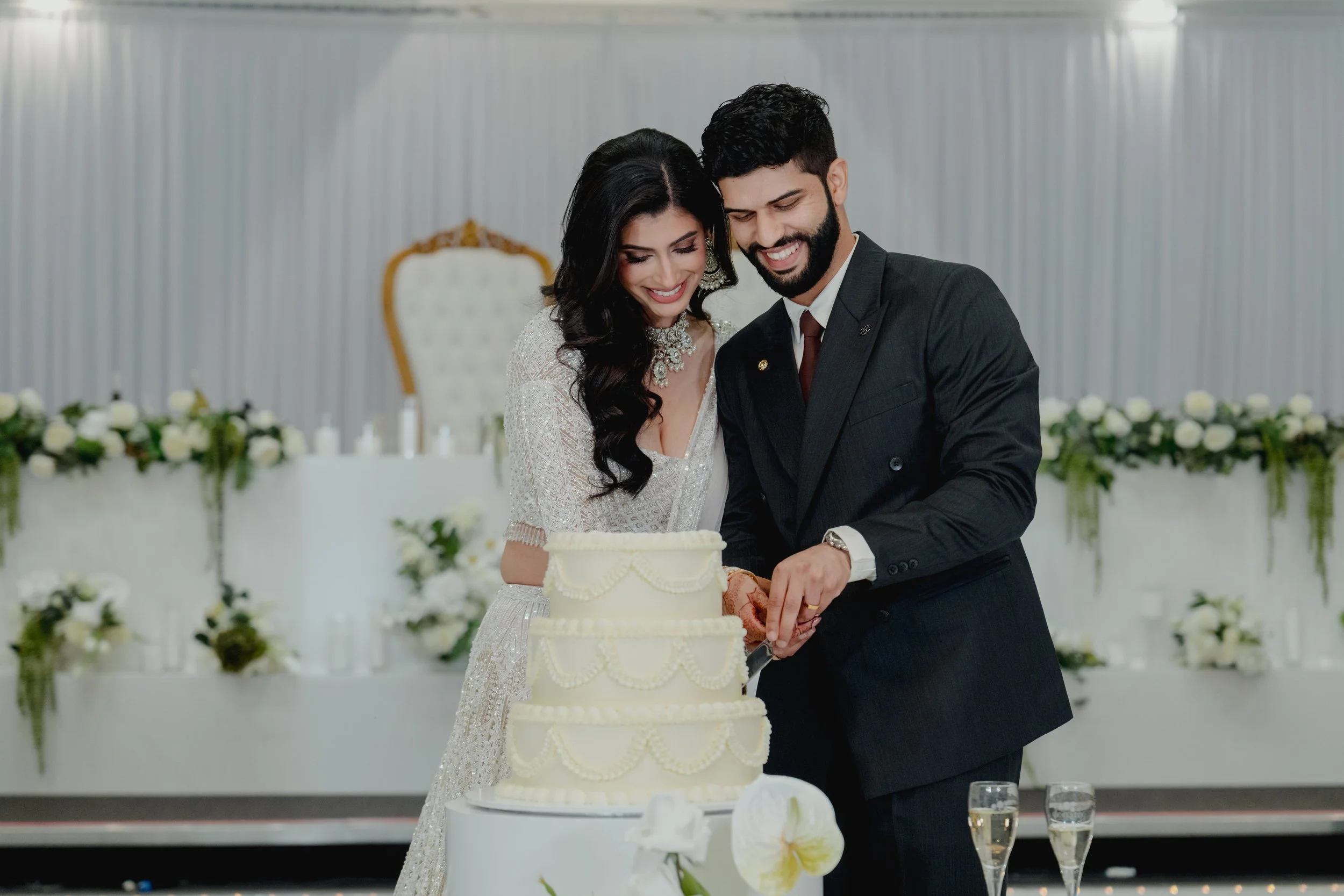 A bride and groom smiling while cutting a wedding cake together at a wedding reception with floral decorations in the background at Donato reception centre in Adelaide