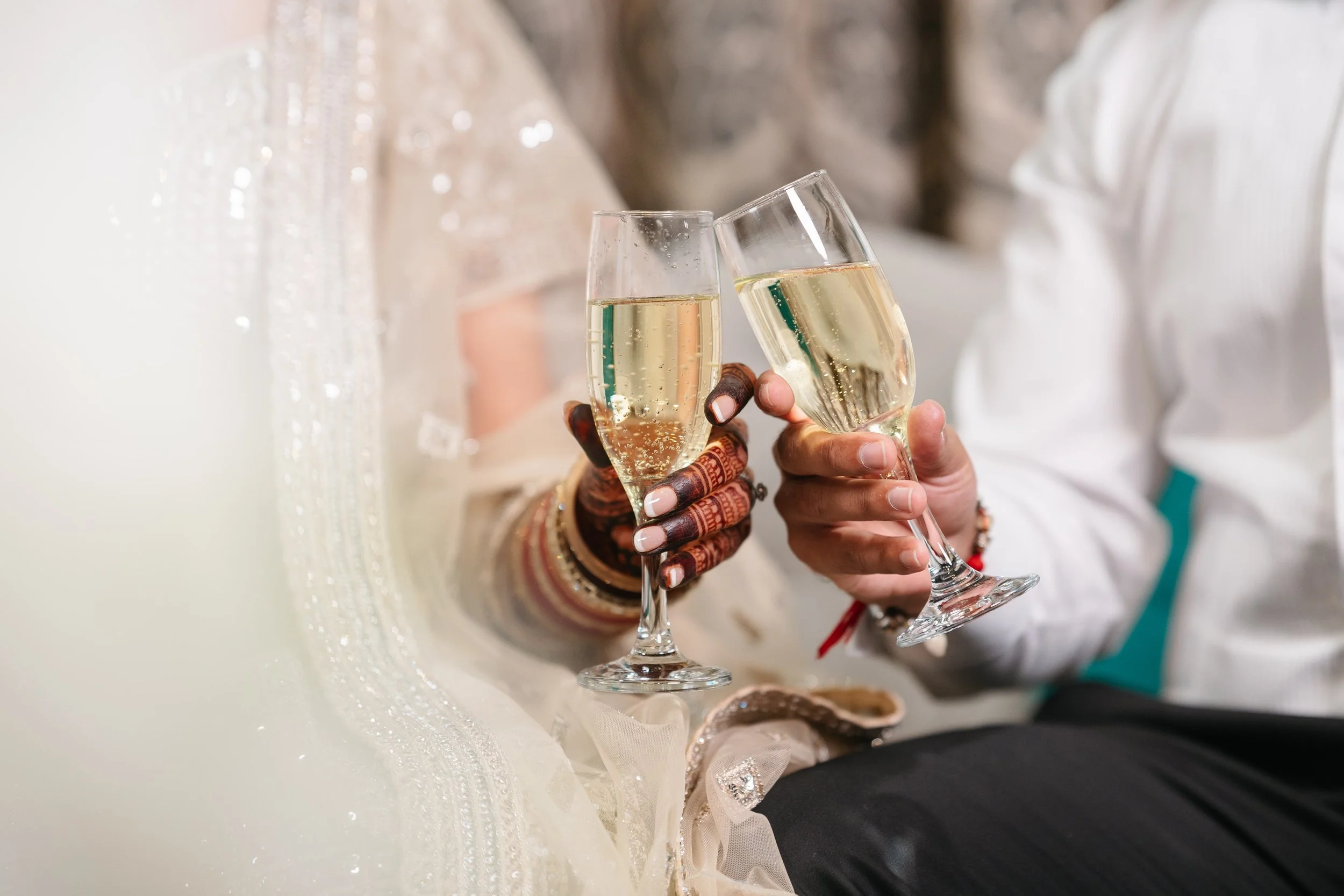 Two women holding champagne glasses clinking at a celebration, with one woman's hennaed hand visible and dressed in festive attire.
