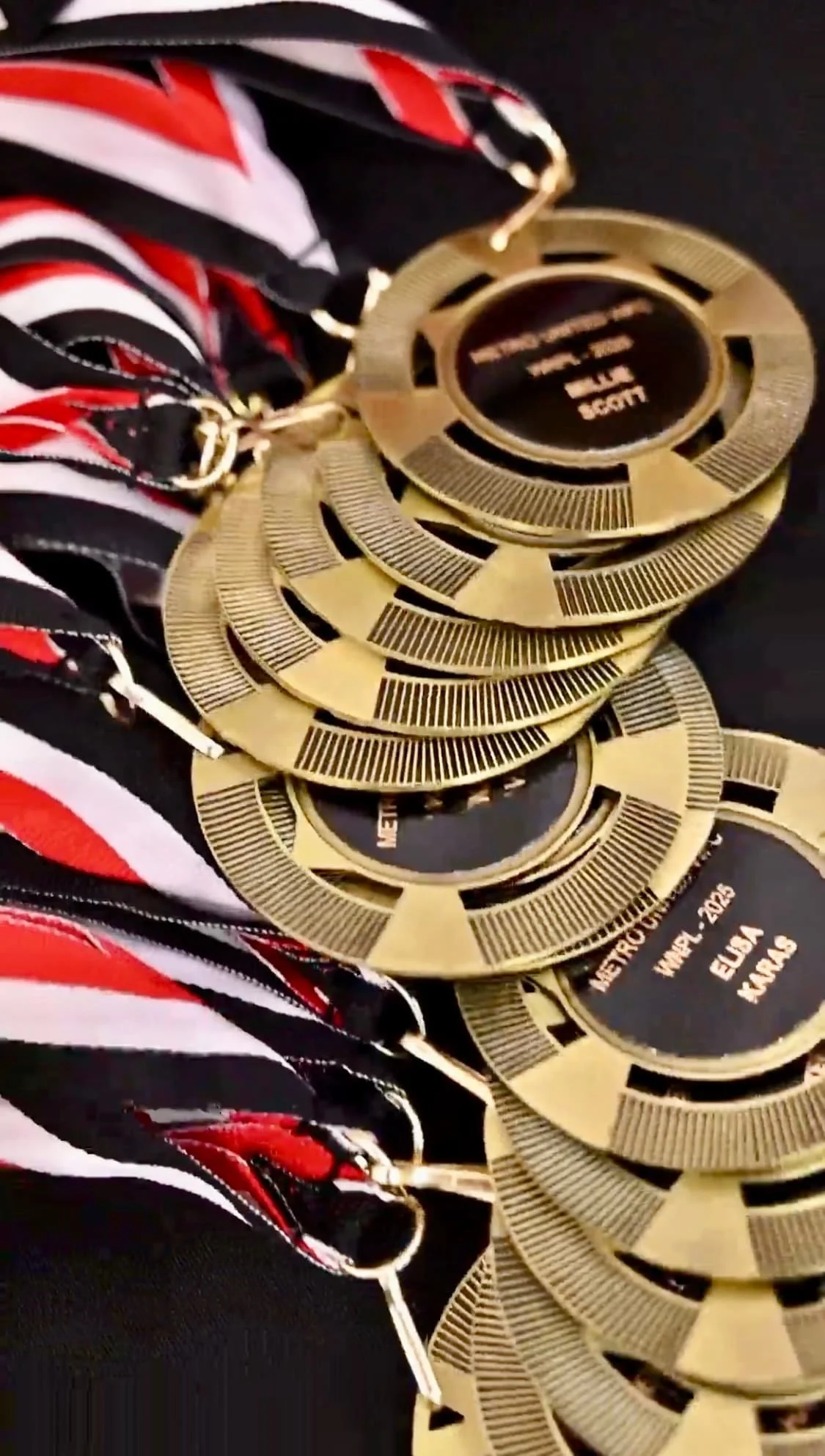 Close-up of medals, with black ribbons decorated with red, white, and black stripes, attached to gold circular medals with engraved text, laying on a dark surface.