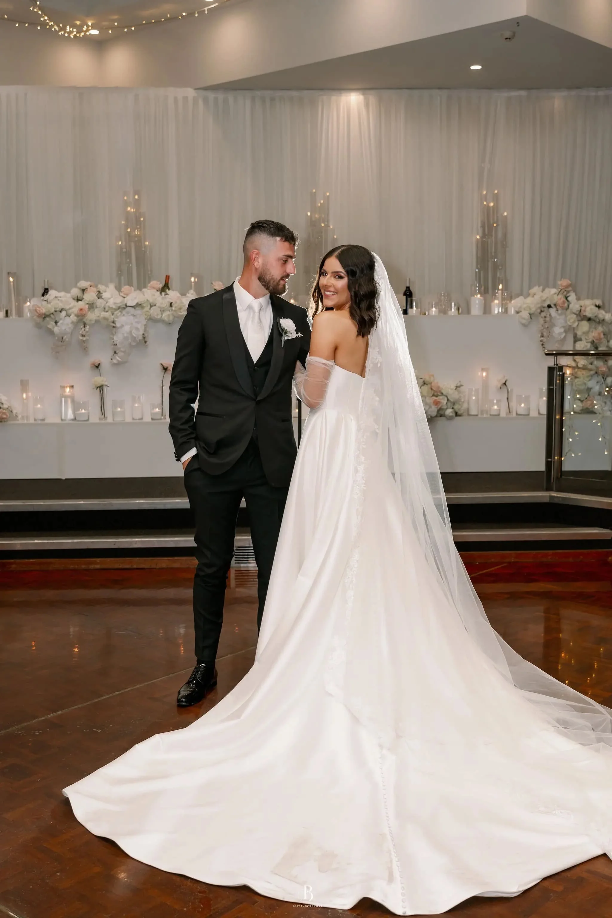 A bride and groom dancing at their wedding reception, with the bride in a white gown and veil, and the groom in a black tuxedo, against a backdrop of floral decorations and candles at Donato reception centre in Adelaide