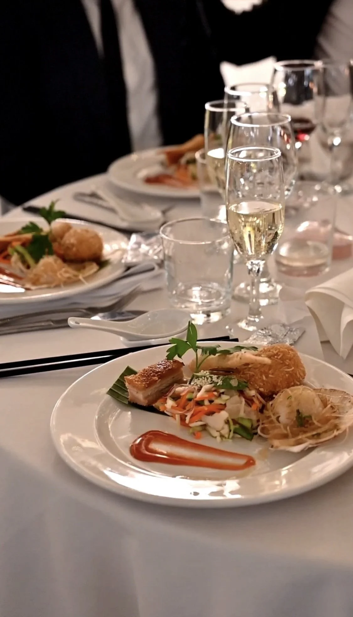 A formal dining table with white tablecloth, set with white plates, silverware, and glassware. There are dinner plates with assorted foods, including fried items, vegetables, and garnishes. Glasses of champagne and red wine are present, and in the ba