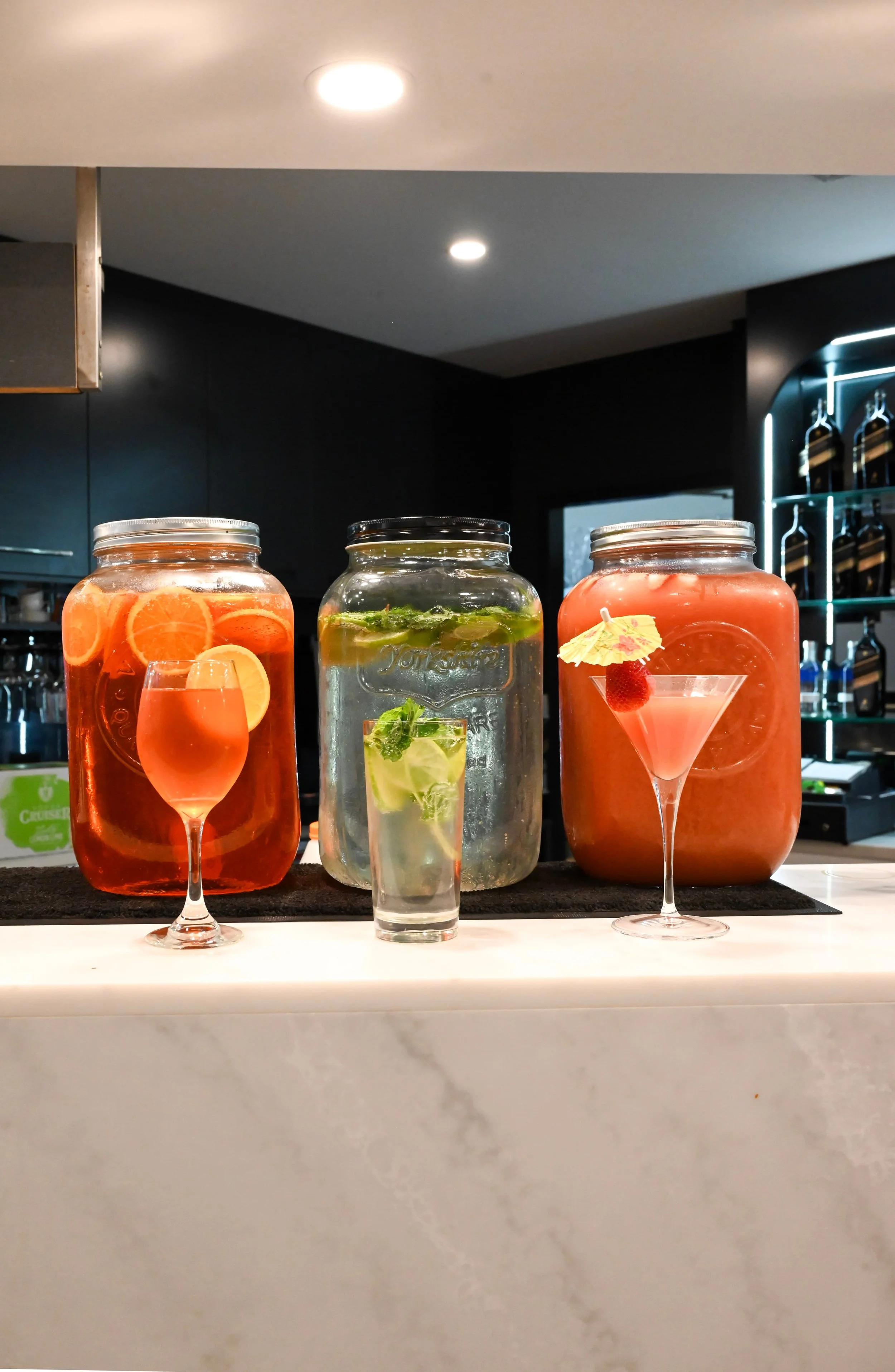 Three jar drinks with fruit garnishes on a bar counter in front of a dark wall and illuminated bottles.