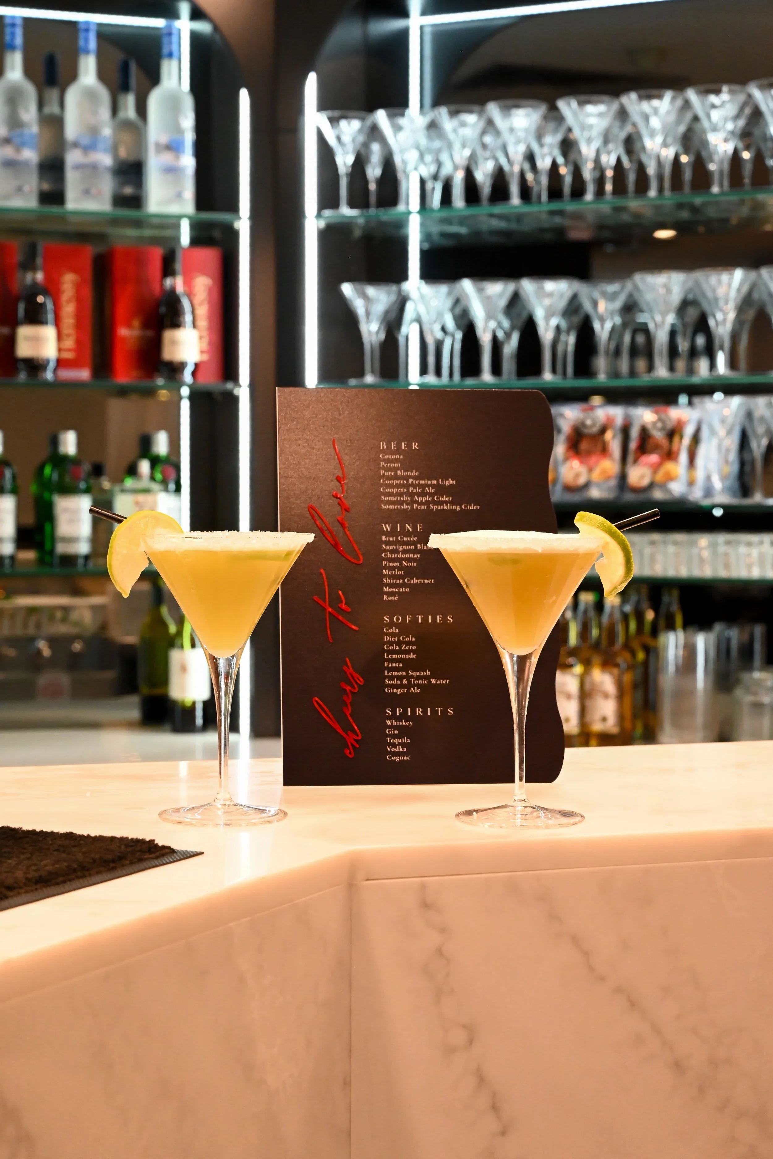Two yellow cocktails garnished with lemon wedges on a bar counter, with a black menu behind them listed with various drink options.