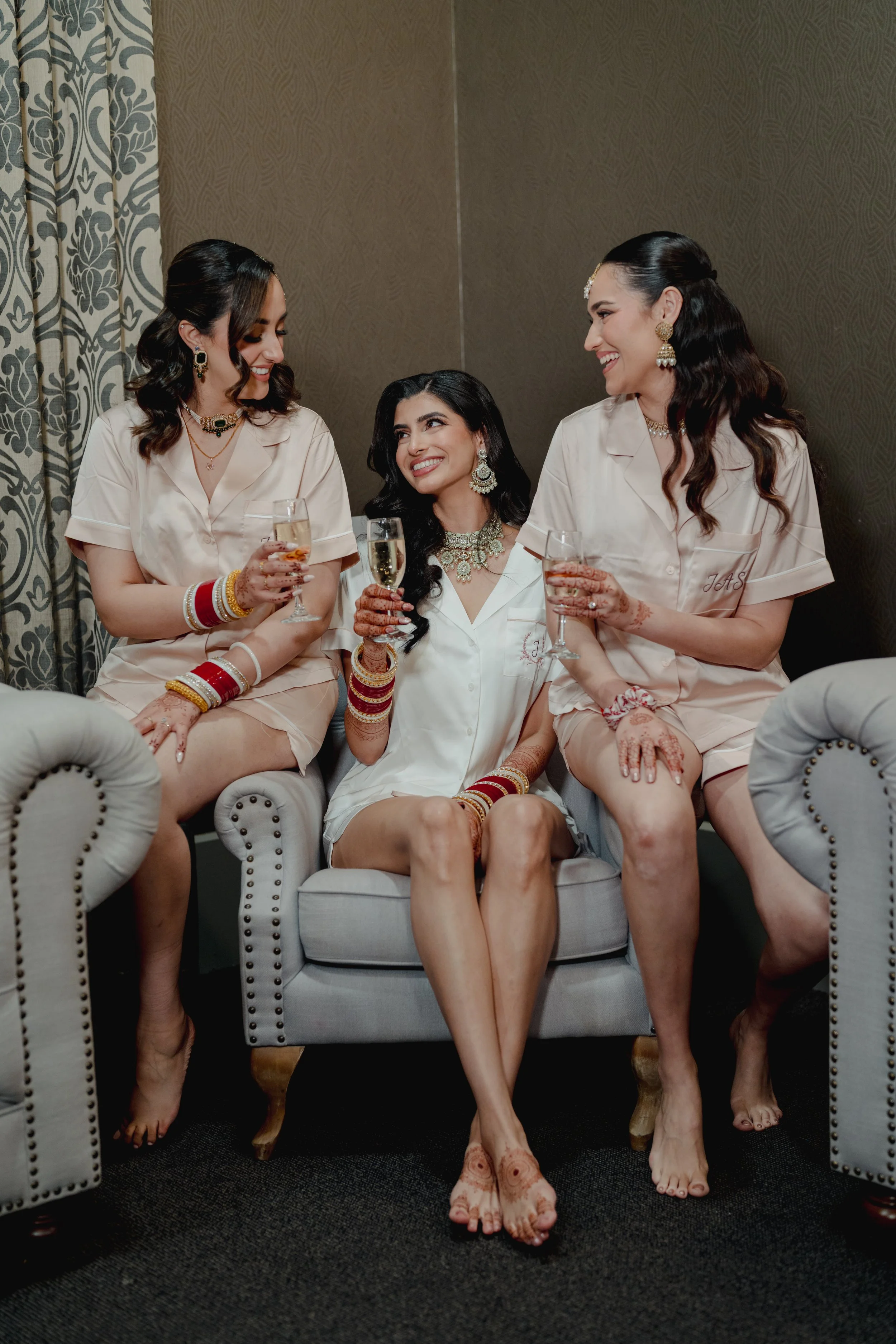 Three women in matching light pink pajamas sitting on a gray sofa, holding glasses of champagne, smiling, and looking at each other. They are wearing traditional jewelry and have henna on their hands and feet, with a decorated background and curtains