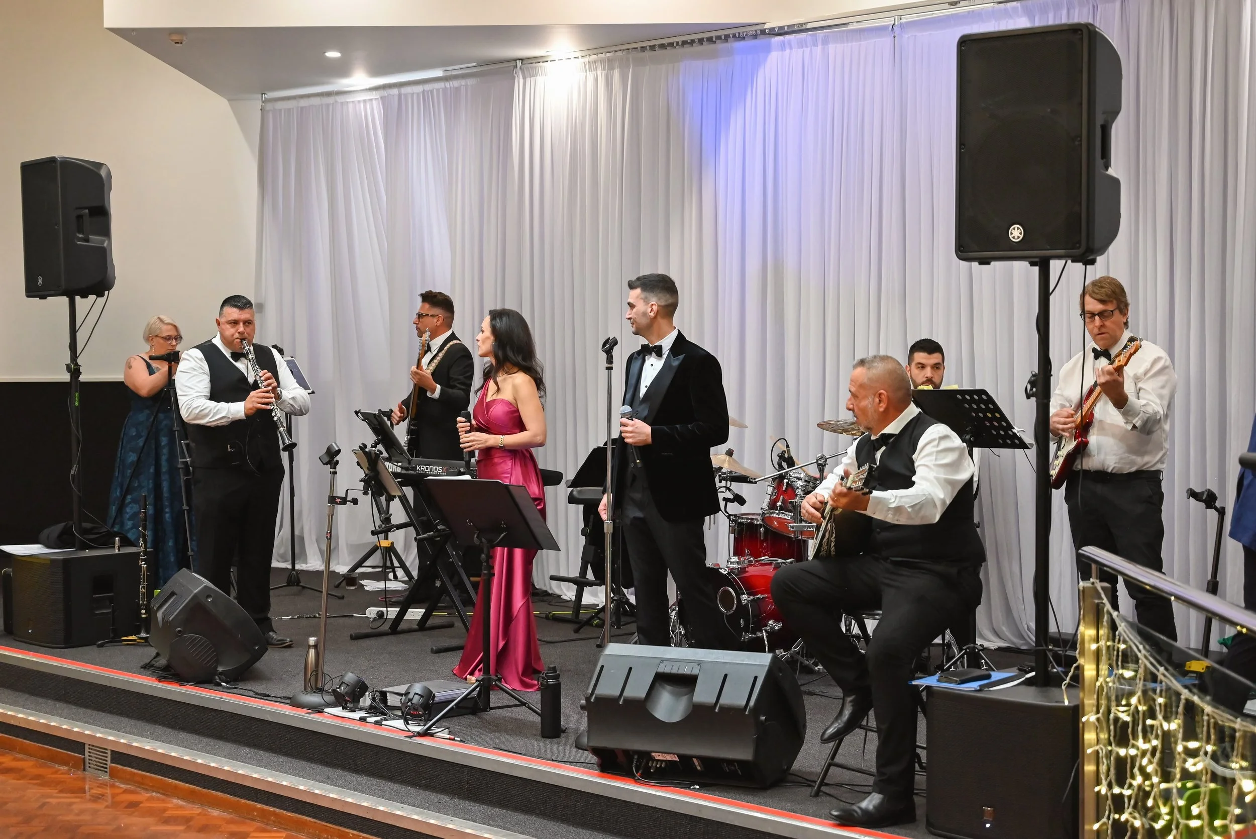 A live band performing on stage at a formal event; the musicians are dressed in tuxedos and dresses, with some playing instruments and singing, and large speakers flank the stage.