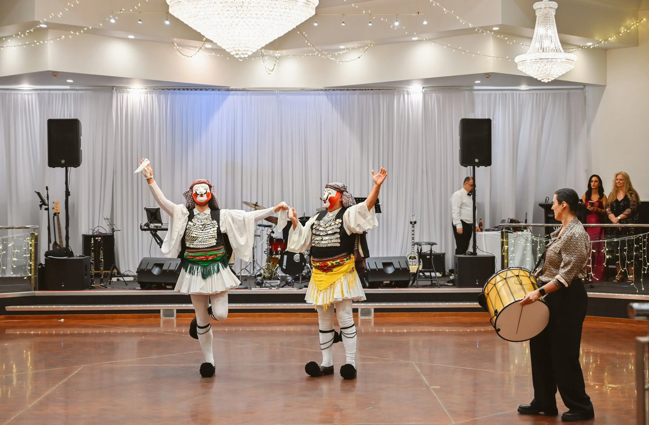 Two performers in traditional Greek costumes dance on a stage, with musicians and singers in the background at a decorated indoor event space.