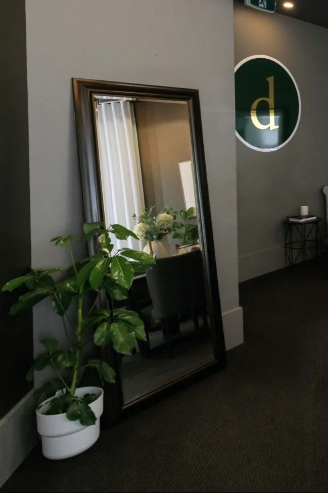 A tall mirror leaning against a gray wall, reflecting a room with curtains and a bouquet of white flowers on a table. A large leafy green plant in a white pot is next to the mirror. To the right, a small black table with a white object on it is visib