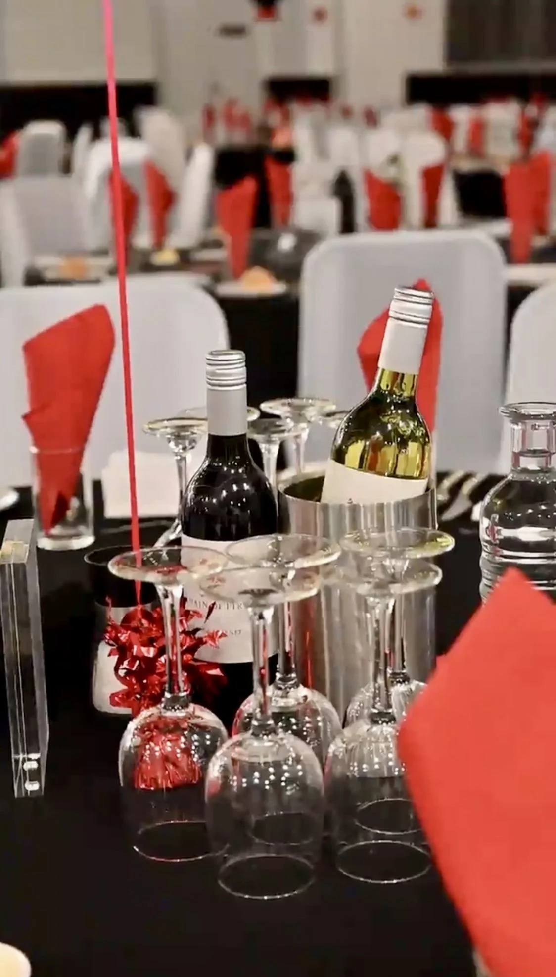 Scene of a decorated table with wine bottles, upside-down wine glasses, and red napkins at a banquet or event.