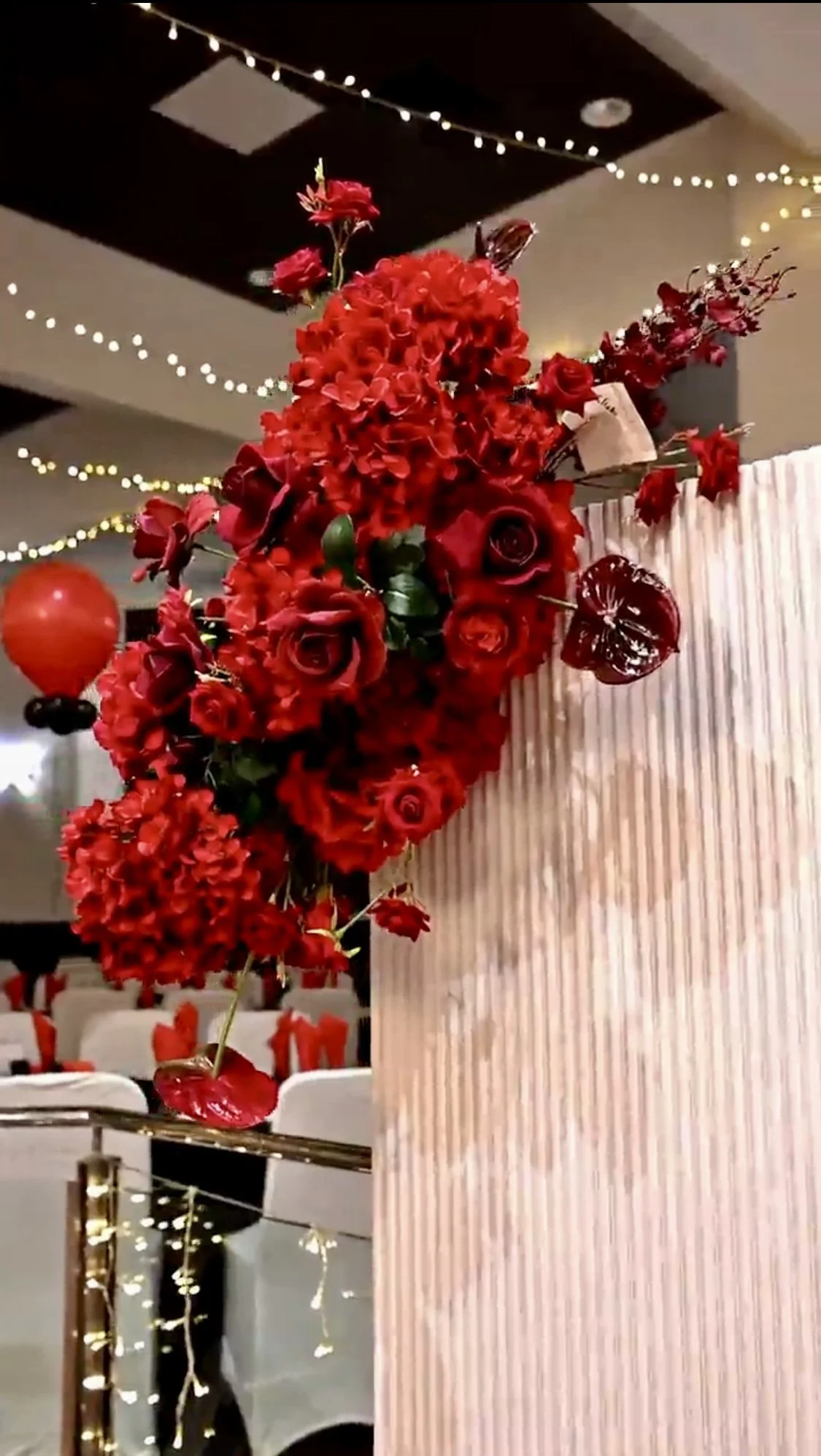 Decorative floral arrangement with red flowers, including roses, orchids, and other blooms, attached to a beige textured wall. String lights are hanging from the ceiling in the background.