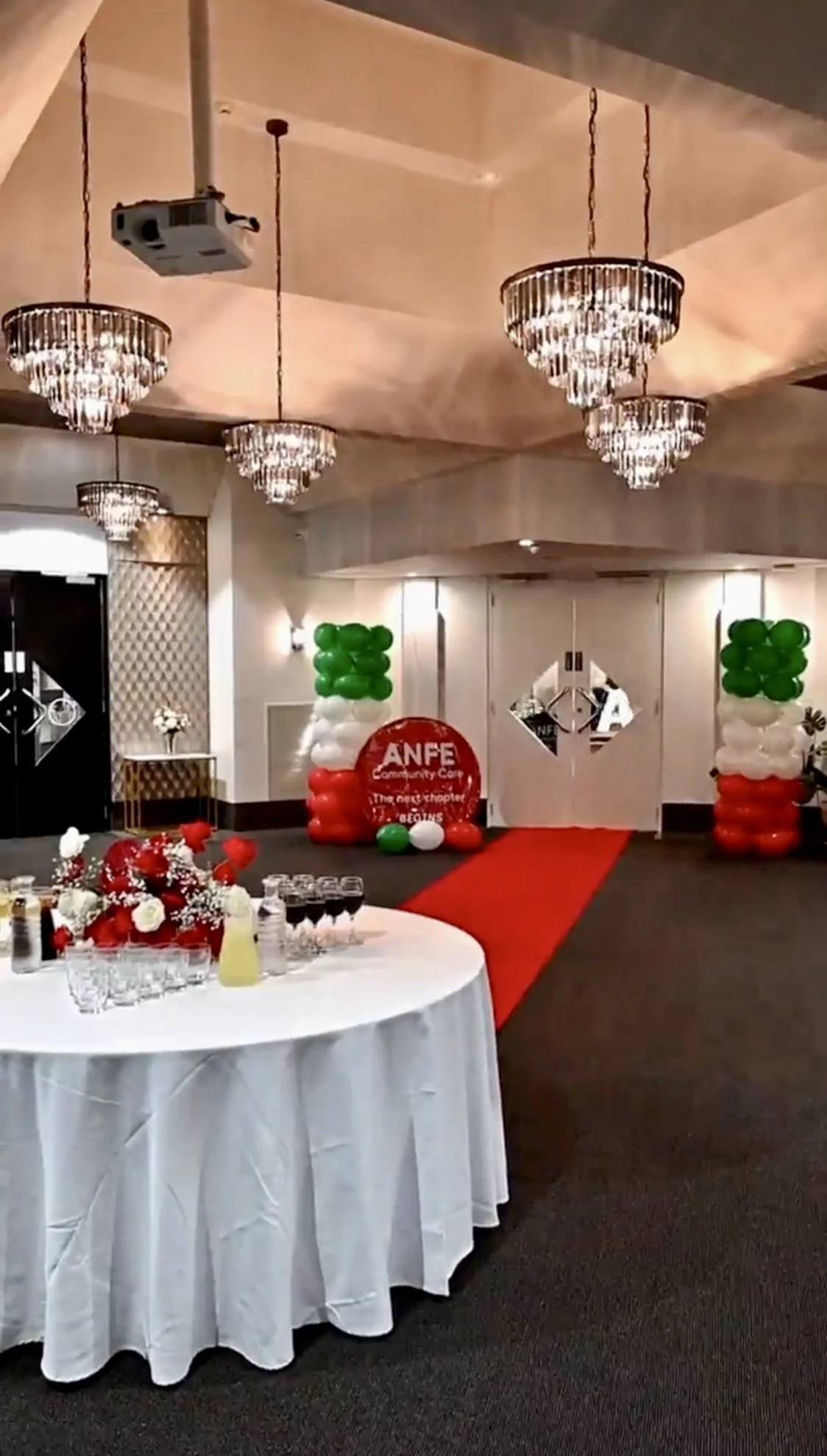 A decorated event hall with multiple chandeliers, a red carpet leading to a door with a sign, balloon arrangements in green, white, and red, a table with flowers, drinks, and glasses, and a white round table with a floral centerpiece.