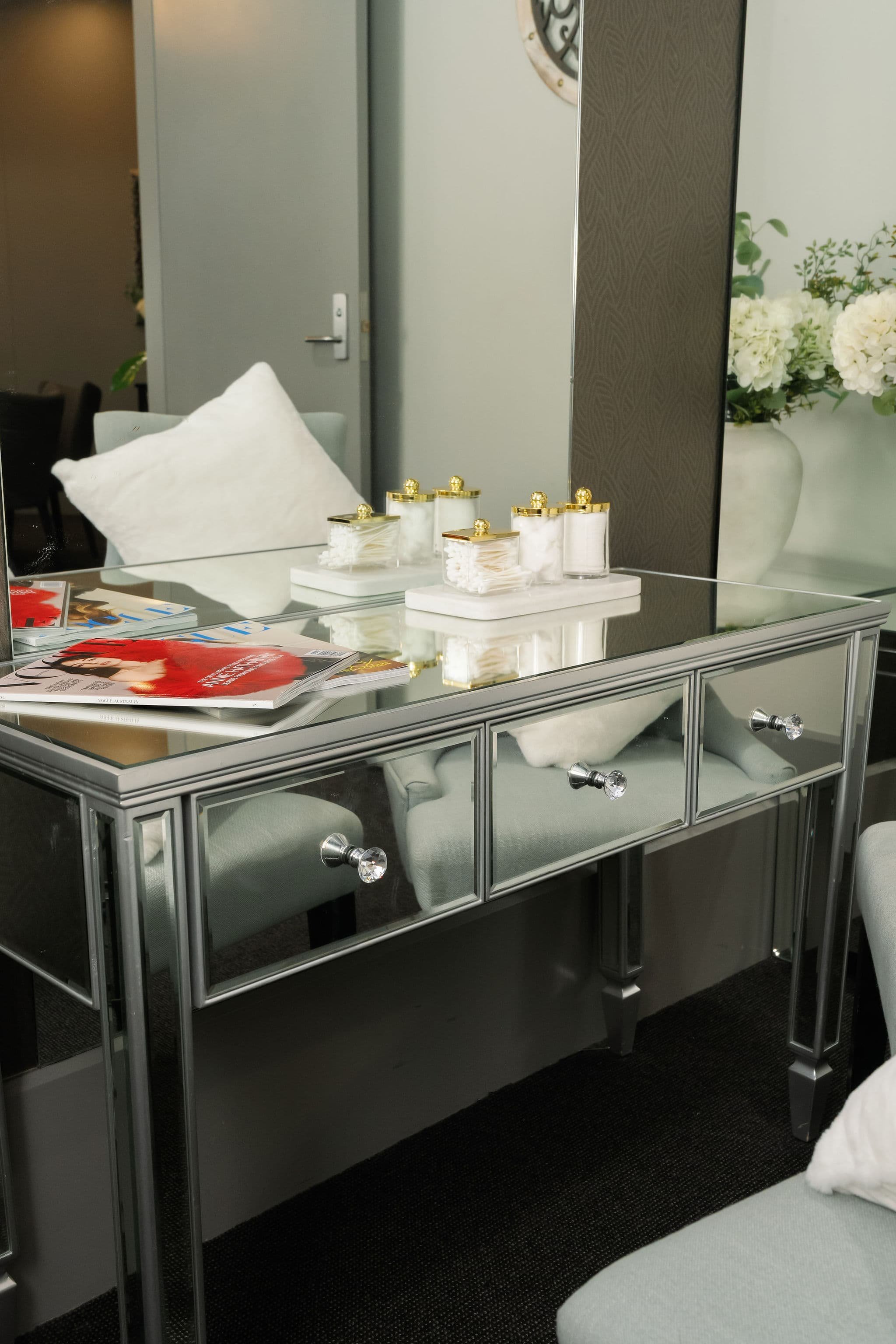 A mirrored display table with magazines on top and various containers with cotton swabs and cotton pads. In the background, there is a white pillow, a beige chair, a door, and a large potted plant with white flowers.