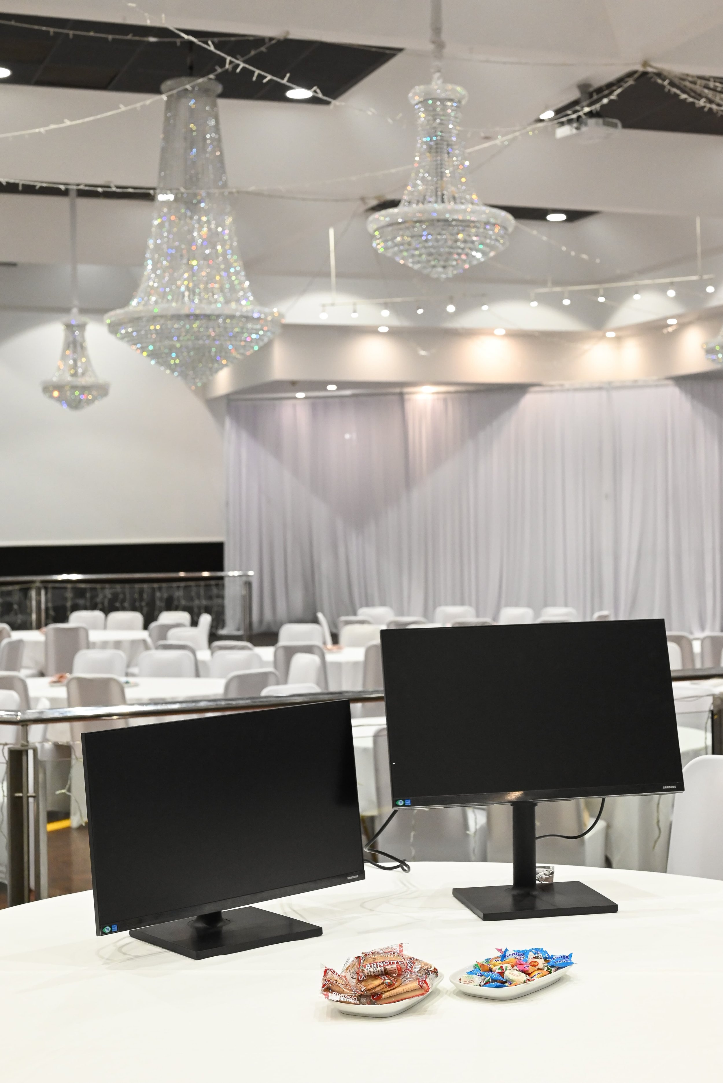 Empty banquet hall with chandeliers, white chairs, and multiple computer monitors on tables; snacks like chips and candy on plates in the foreground.