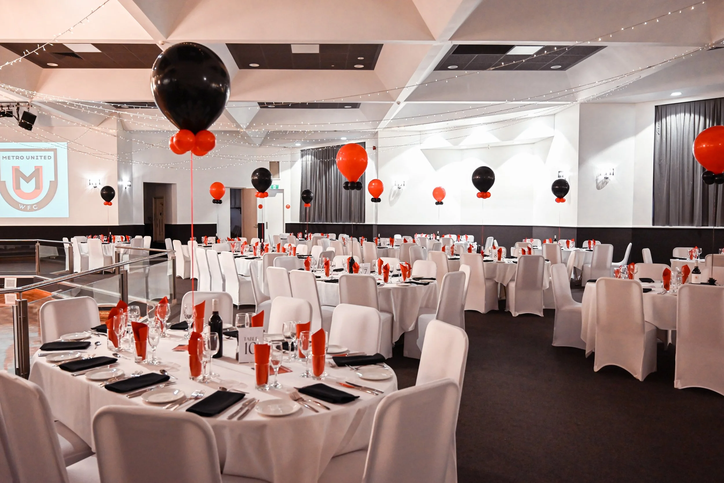 A banquet hall decorated with black and orange balloons, fairy lights, and set tables with black napkins and red folded paper accents, prepared for an event.