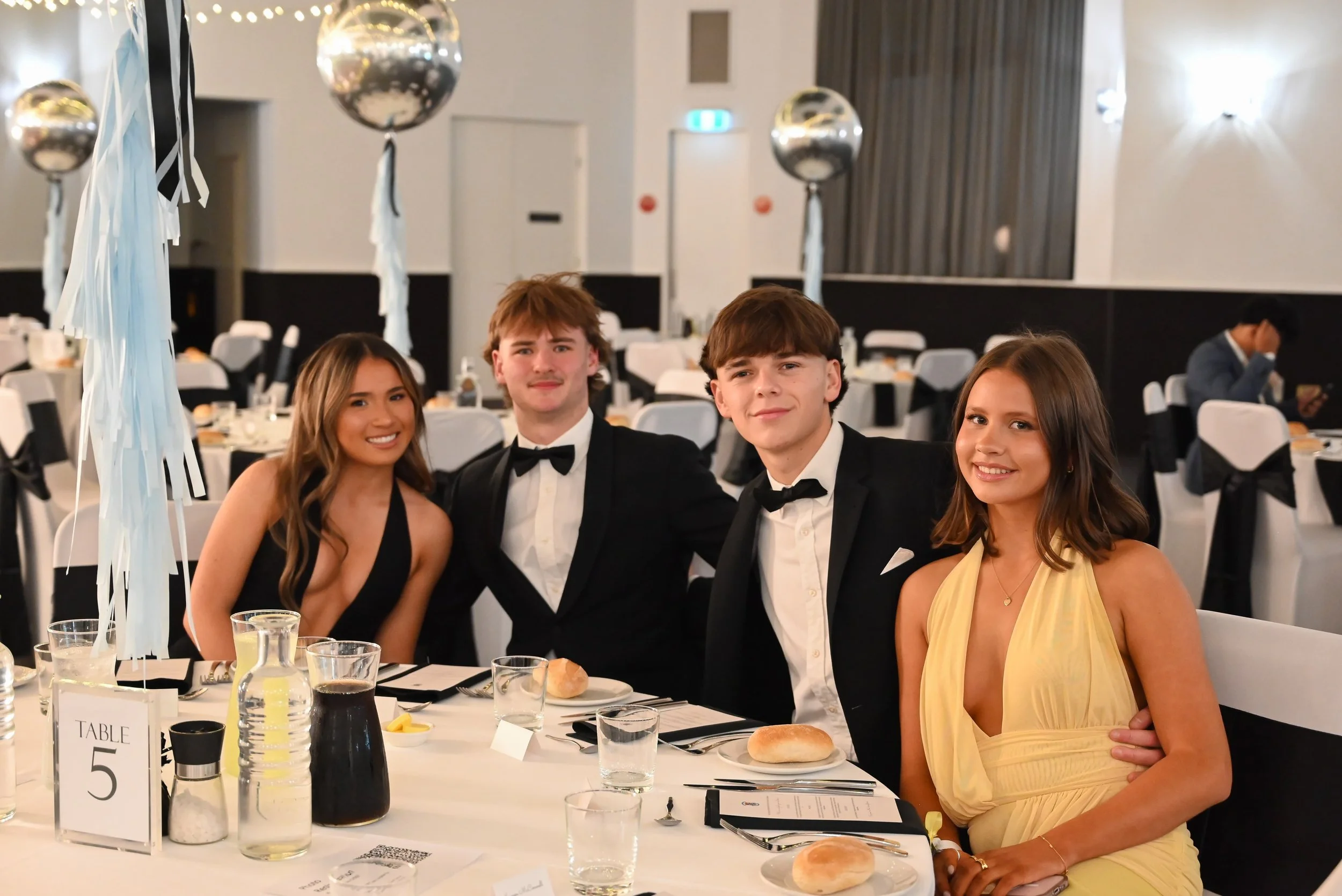 Four young adults dressed in formal attire sitting at a banquet table in a decorated event hall. The table has bottles, glasses, and a sign indicating Table 5. Silver balloons with white ribbons are part of the decoration. Two women and two men are s