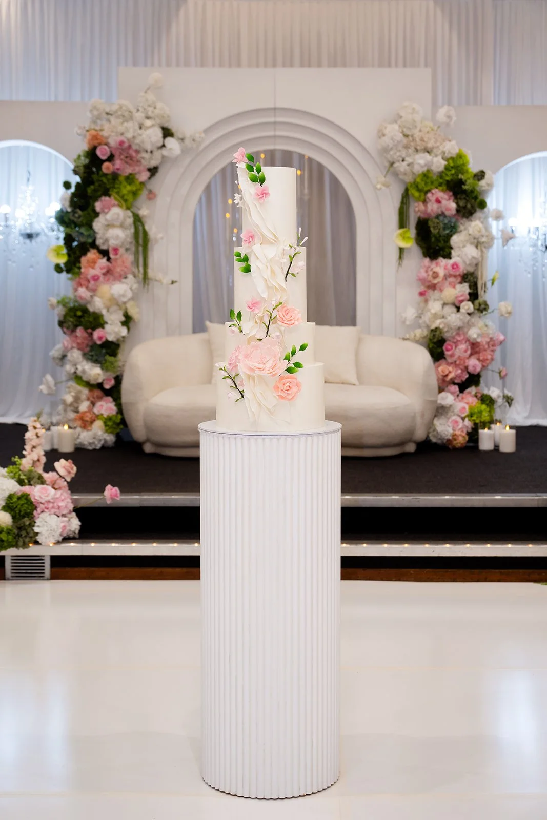 Elegant wedding cake with pink and white flowers, displayed on a white, fluted pedestal in front of a floral and white drapery backdrop with a loveseat and floral arch  at Donato the venue