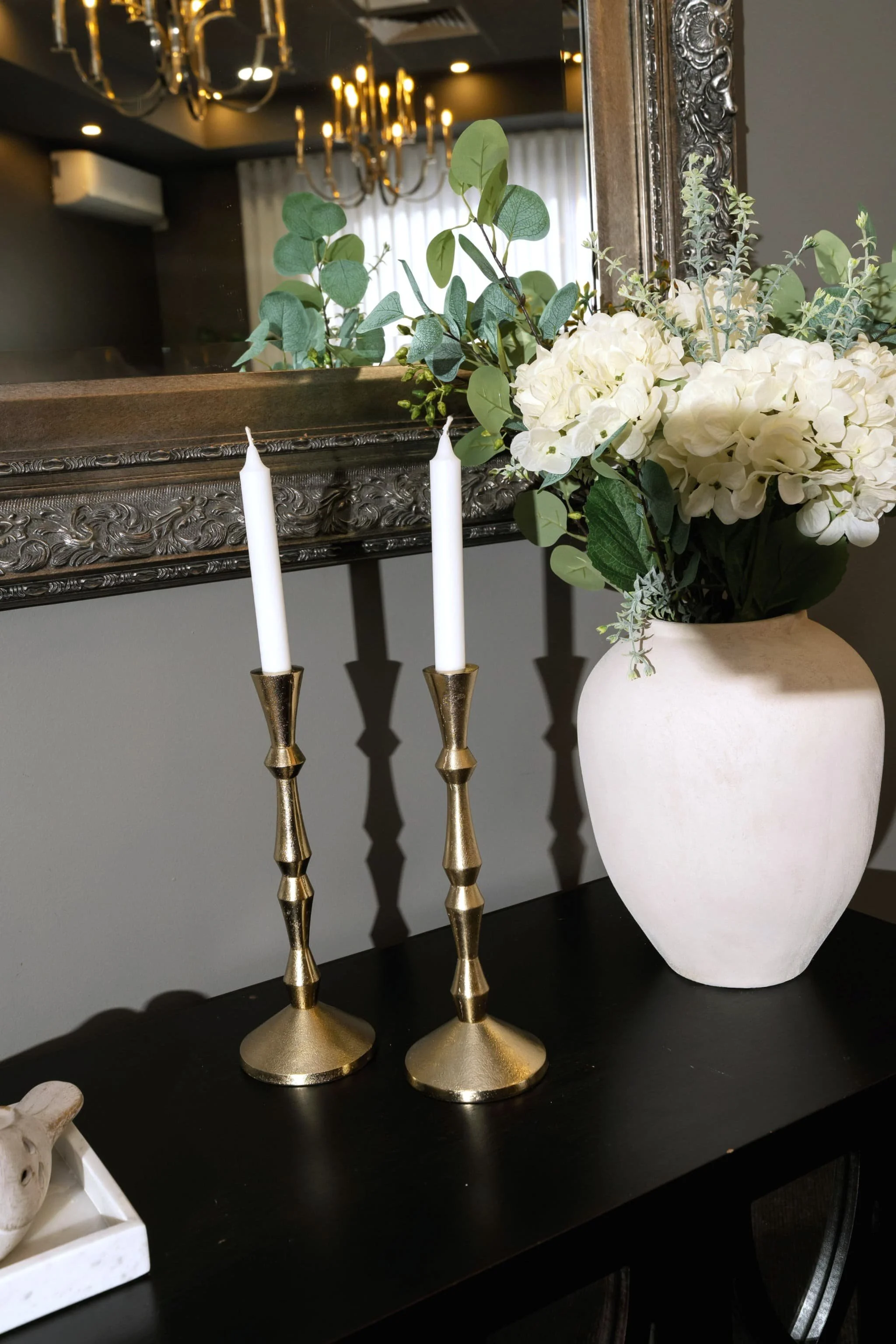 Decorative table setting with a large white ceramic vase filled with white hydrangea flowers and greenery, and gold candlesticks with white tapered candles on a black table. A mirror and chandelier are visible in the background.