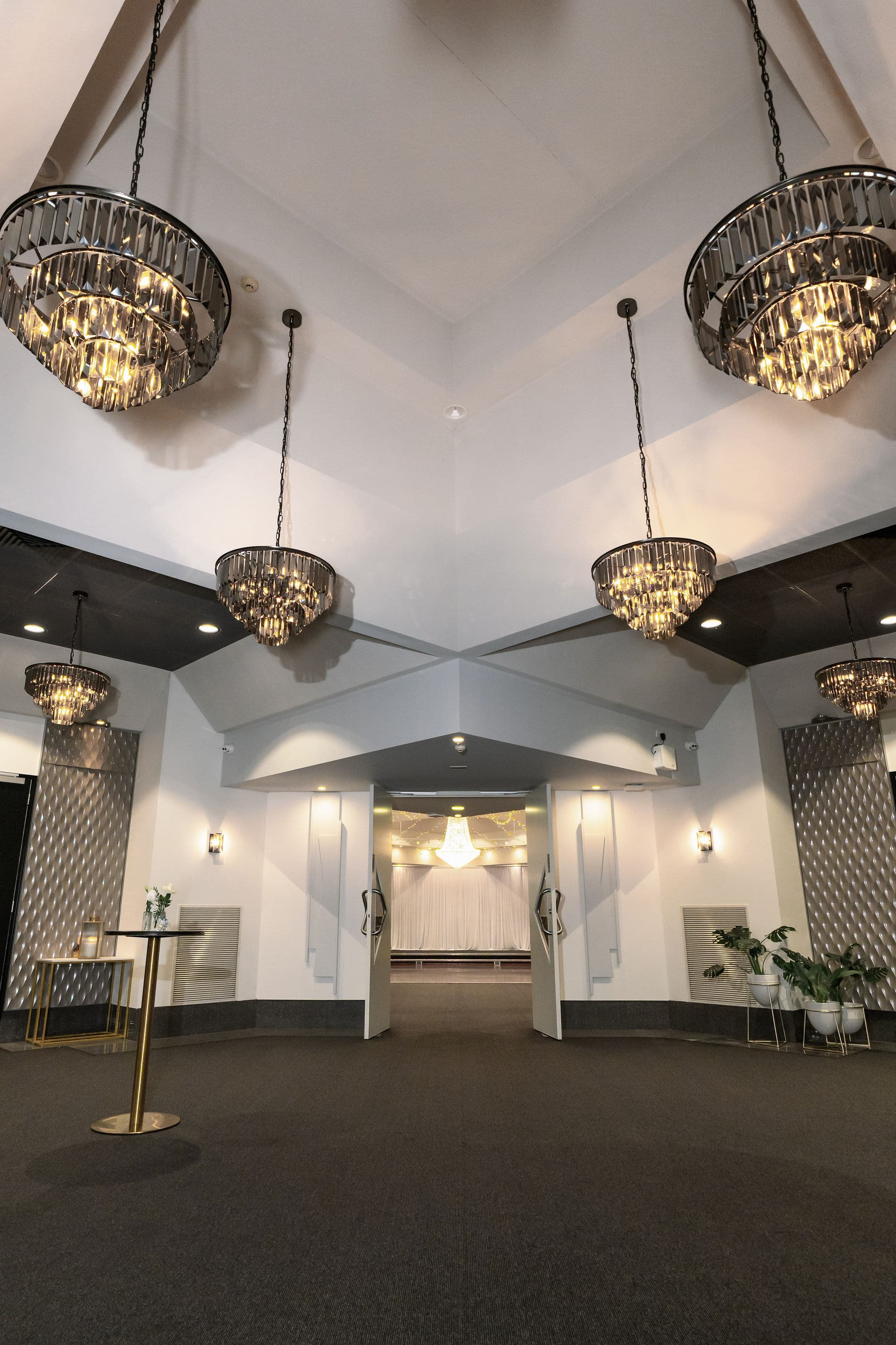 Elegant indoor space with four large chandeliers hanging from the white ceiling, white walls, potted plants, a small table with a flower arrangement, and a doorway with a chandelier in the background.