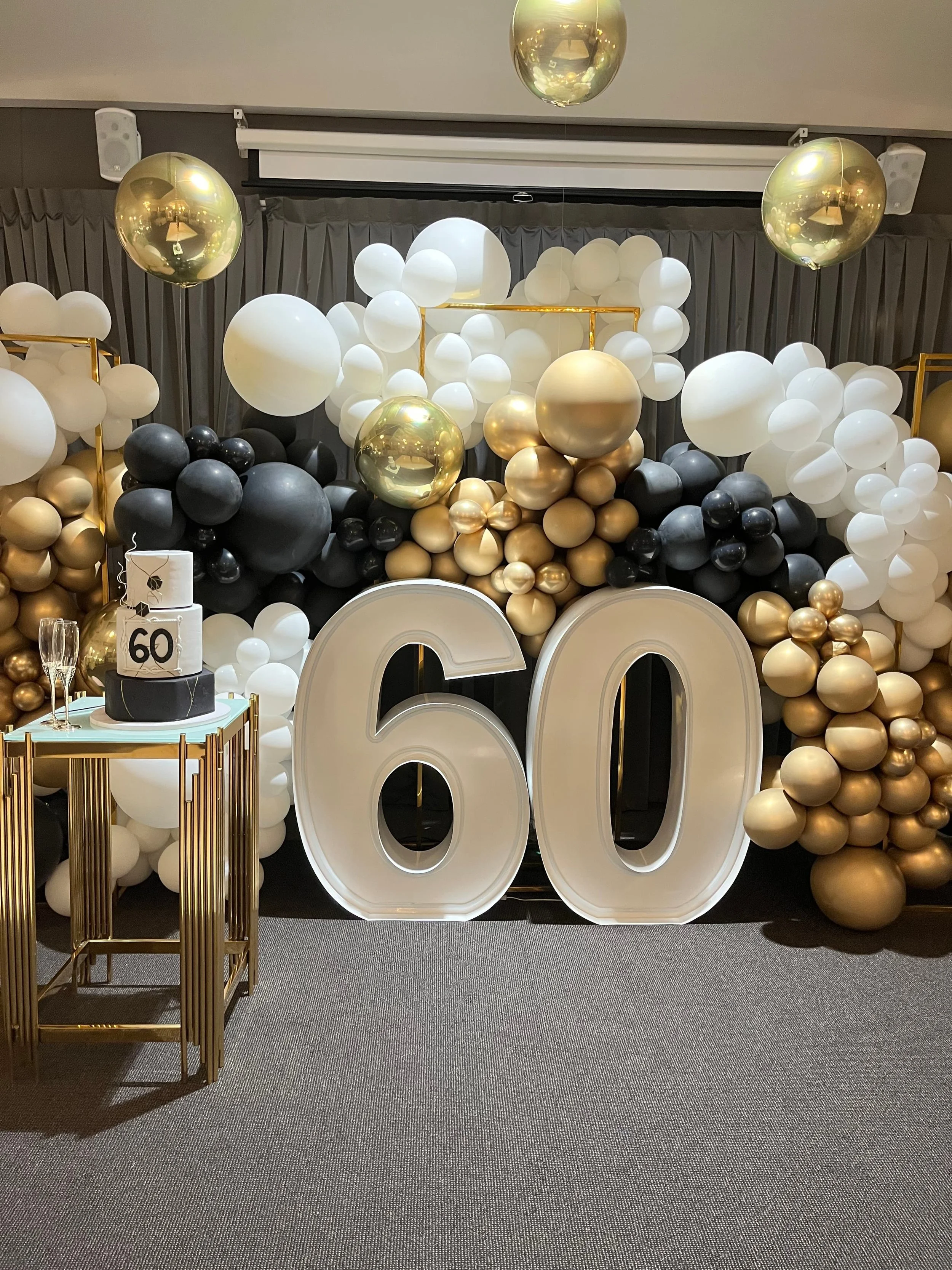 Decorative balloon arrangement with large white, black, and gold balloons and giant number '60' cutouts, celebrating a 60th birthday or milestone.