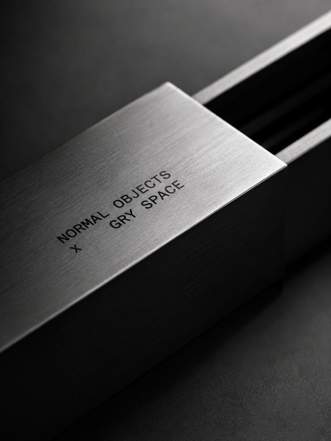 Close-up of a metallic box labeled 'Normal Objects x Grey Space' with a sleek, brushed metal finish on a dark surface.