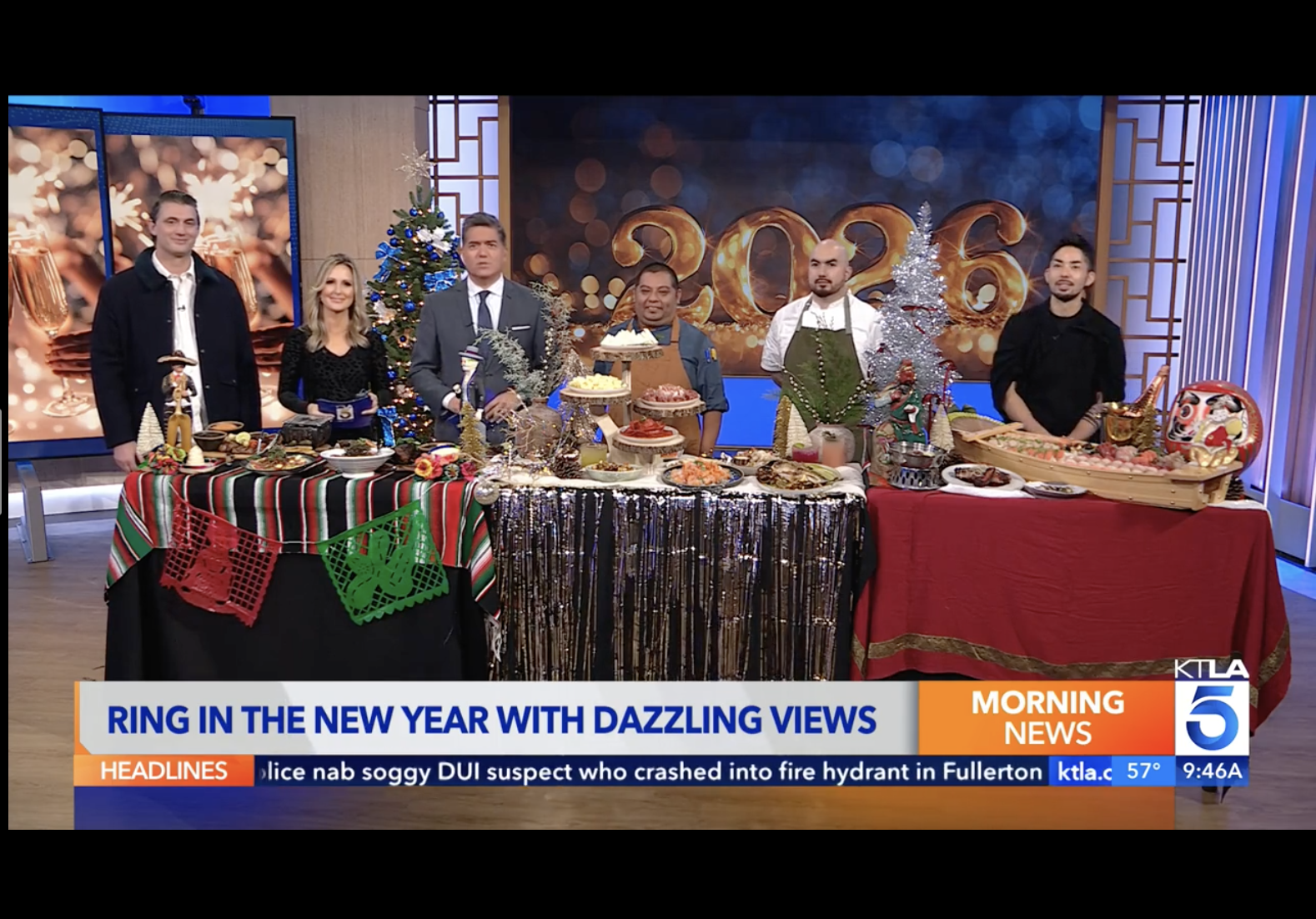Television news segment with a group of people standing behind tables with festive food and decorations, celebrating New Year's with a digital backdrop reading '2026'.