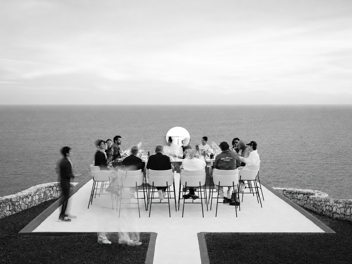 A group of people dining at a table outdoors on a terrace overlooking the ocean, with a cloudy sky above.