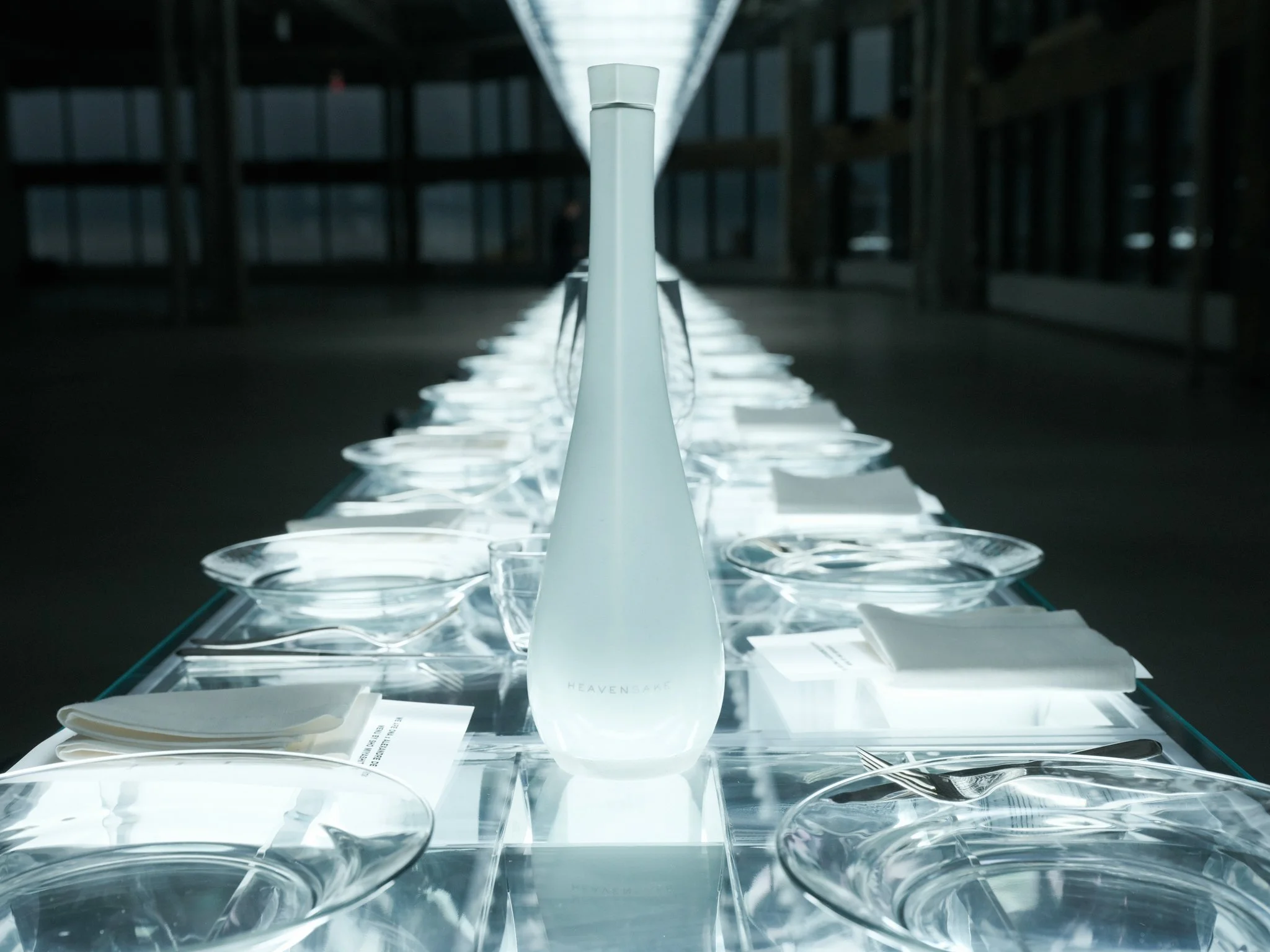 A display of white, transparent, and light pink glassware and ceramic objects arranged on a reflective surface in a dark, modern exhibition space. The objects are illuminated from above, creating reflections and highlights.