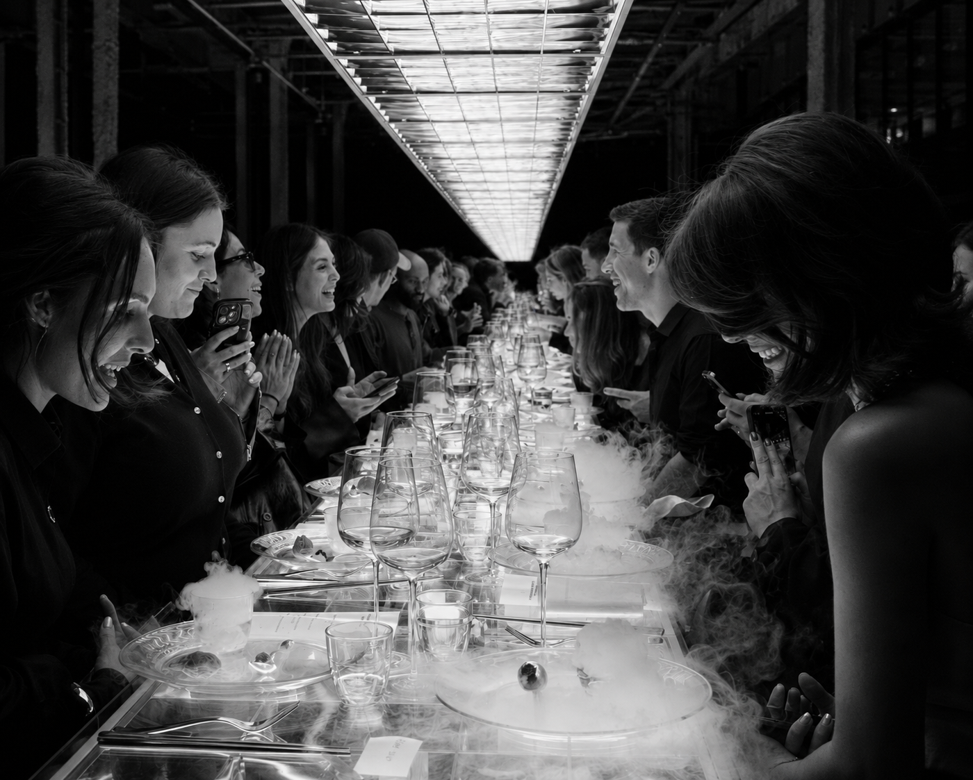 Black and white photo of a large group of people sitting at a long table, smiling and taking pictures of a dessert with dry ice smoke in front of the camera