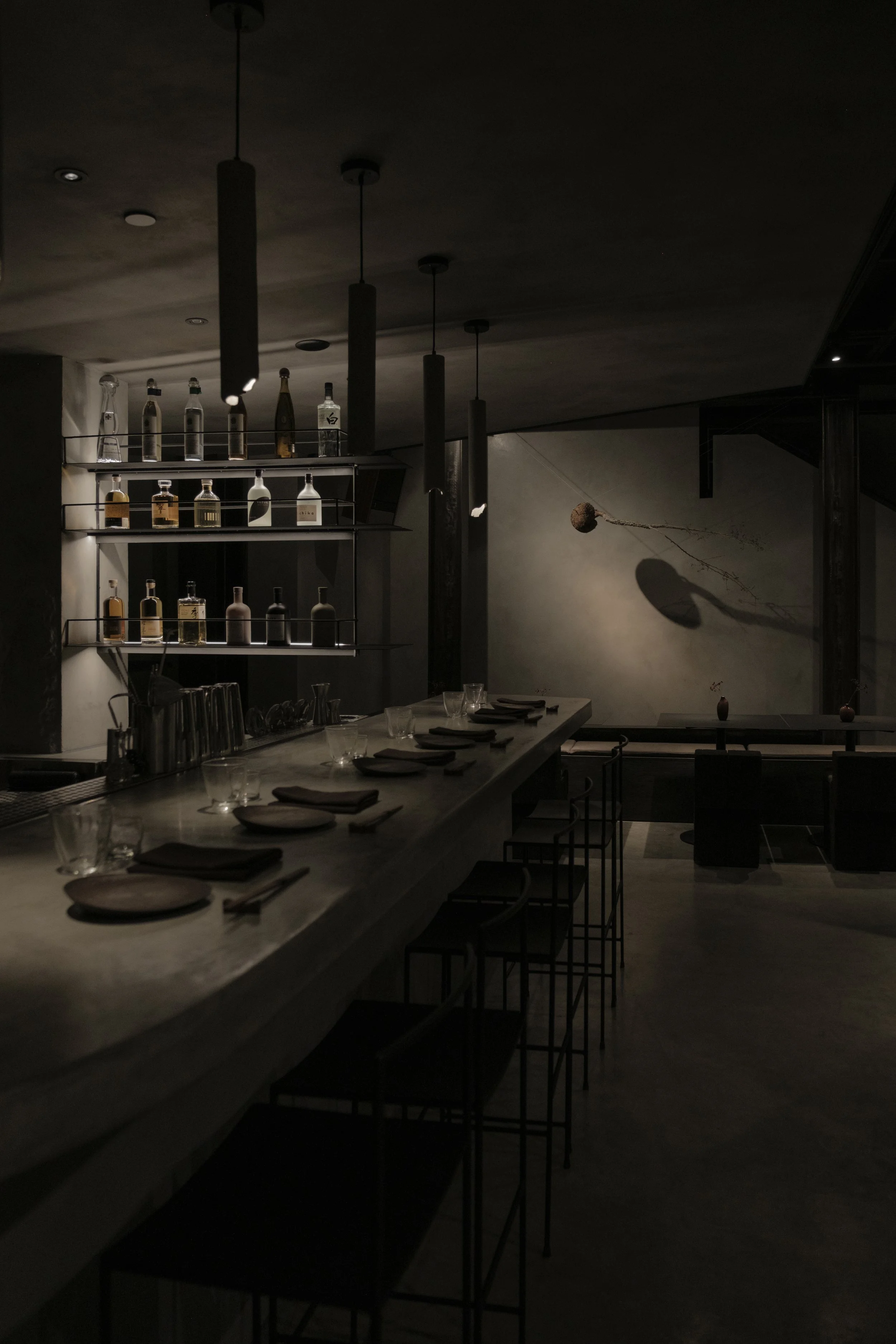 A dimly lit bar with a concrete countertop, glasses, and neatly folded napkins, with shelves of bottles and hanging pendant lights, and a wall decorated with a shadow of a hanging object.