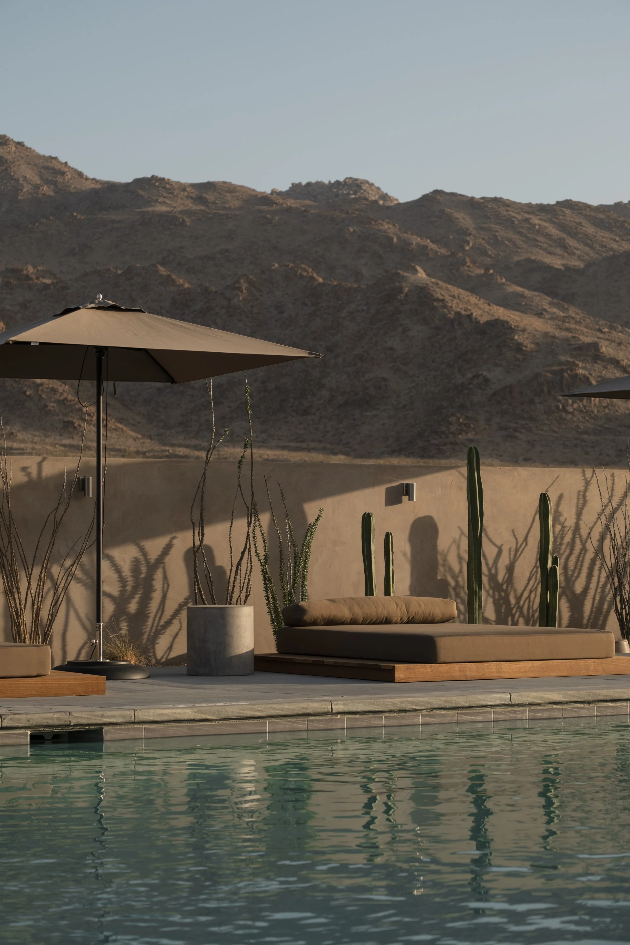 A desert landscape with mountains in the background and a pool in the foreground. There are patio umbrellas, a cushioned lounge chair, and potted cacti near a beige wall.