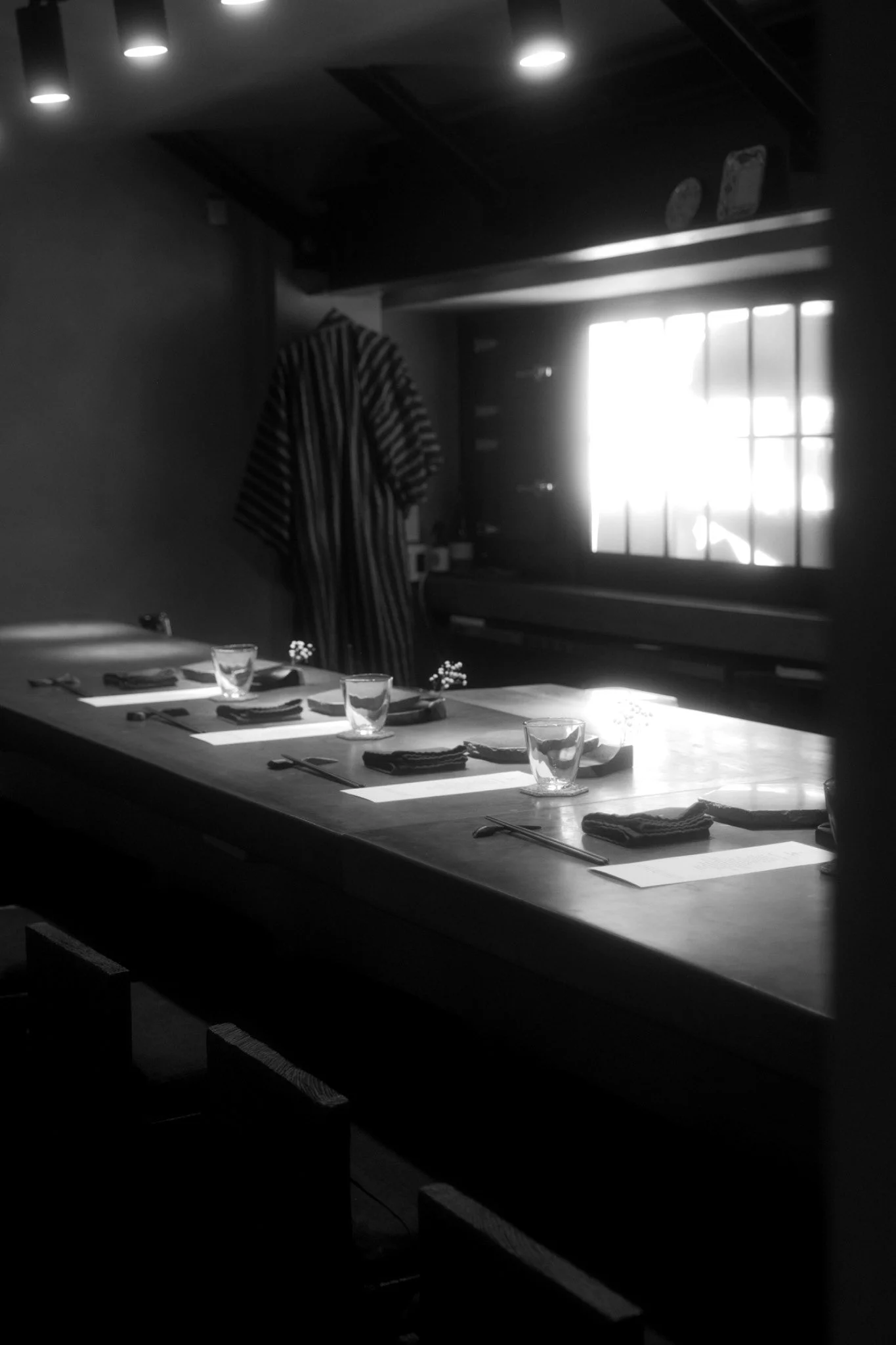 A dining table set with glasses, napkins, and utensils, illuminated by sunlight coming through a window with multiple panes, in a room with a dark ceiling and a patterned curtain on the wall.