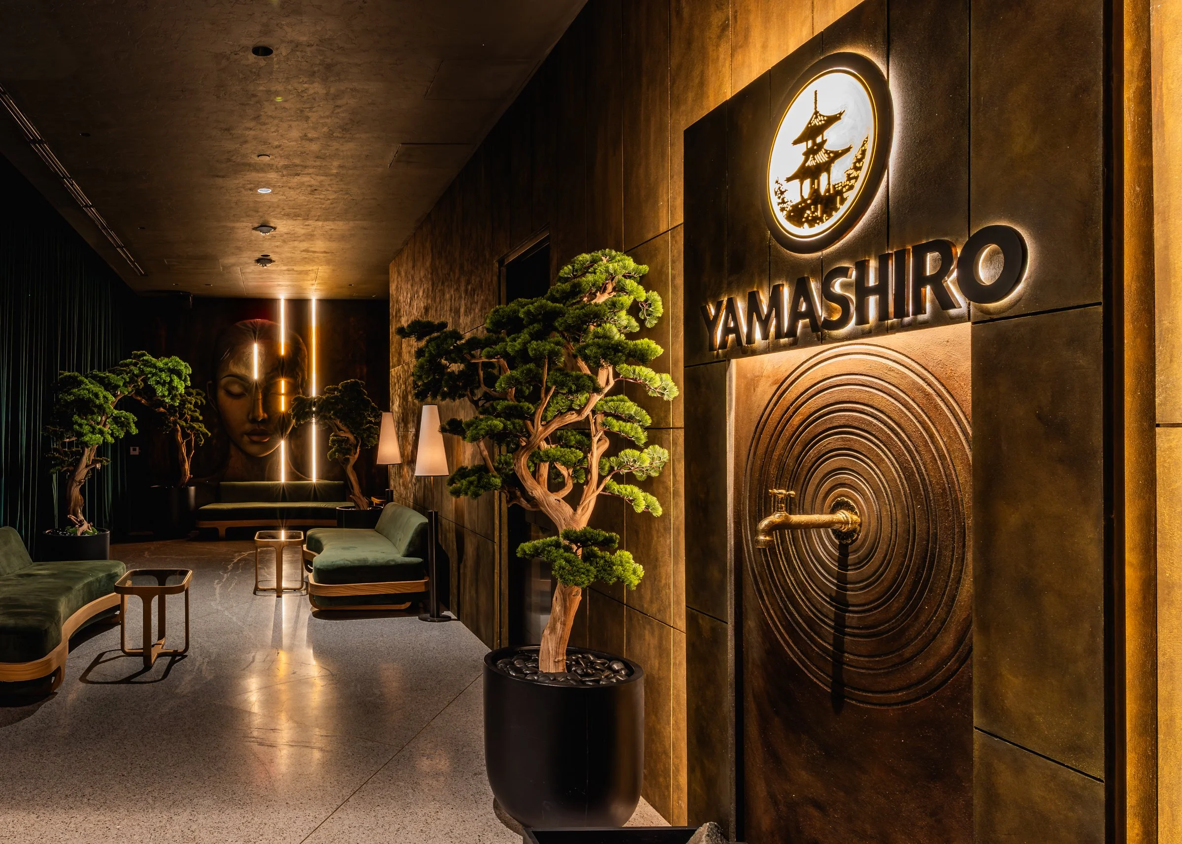 Lobby area with green velvet seating, bonsai trees in black pots, wall art of Buddha, illuminated signage with a pagoda and the word 'YAMASHIRO.'