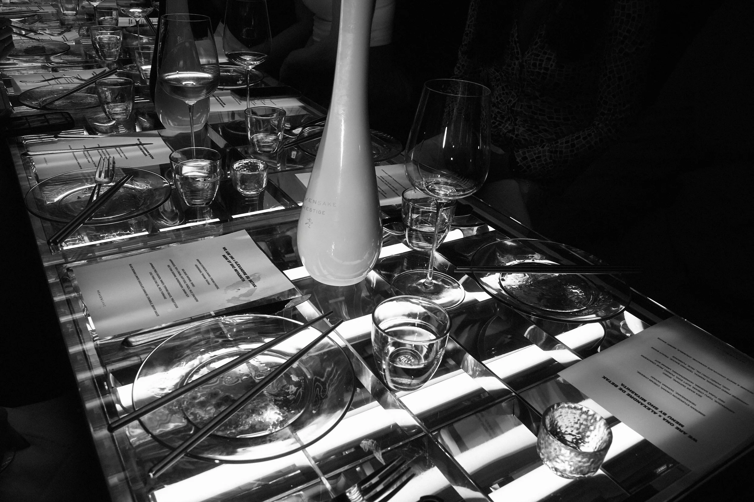 A dining table set with various glasses, plates, and chopsticks, illuminated from underneath. There is a white bottle in the center of the table, and menus are visible on the table in front of some places. The setting appears to be in a restaurant.