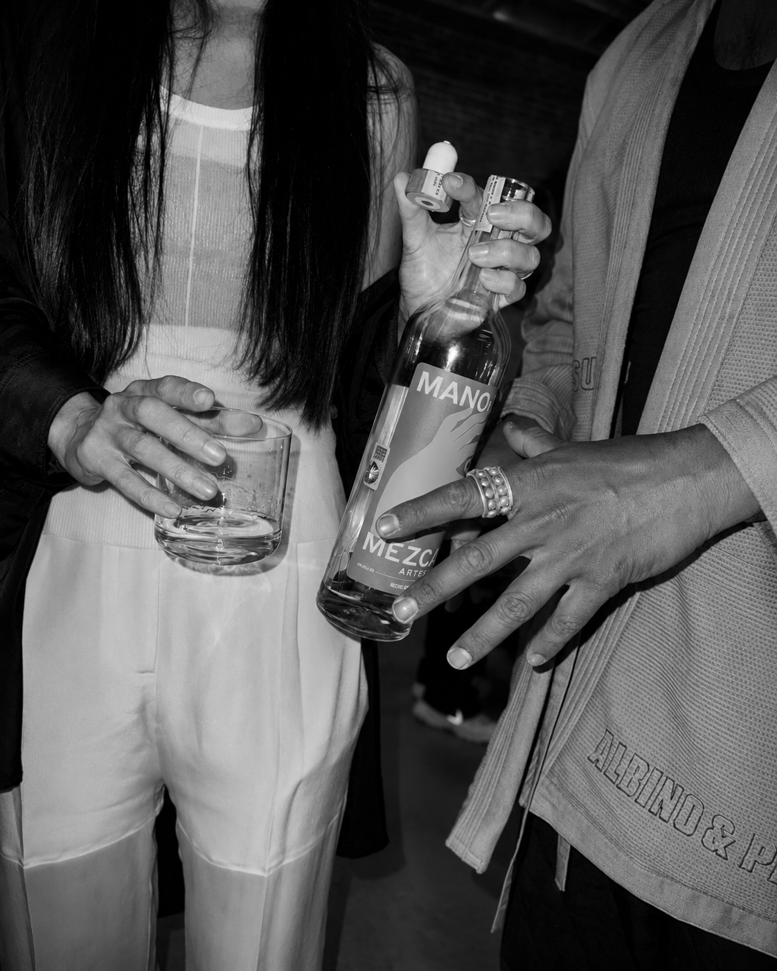 Two women are holding a bottle of mezcal and a small glass, with one woman pouring a shot while the other holds an empty glass.