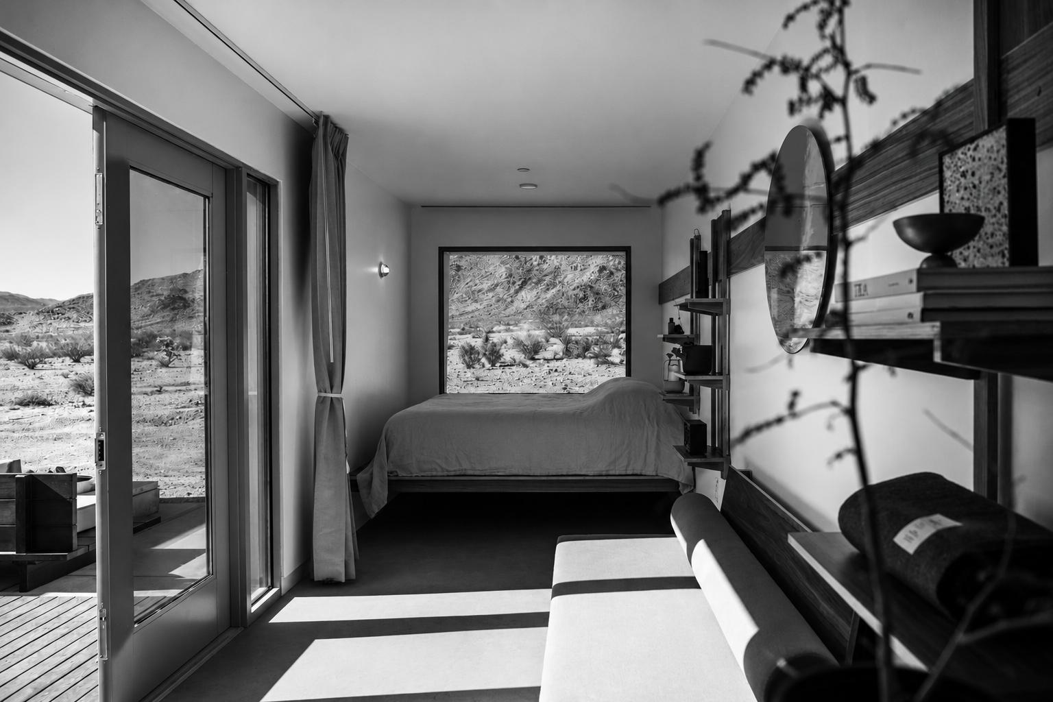 A modern bedroom with large desk and shelving, view of desert landscape through window, and sliding glass door leading outside, black and white photograph.