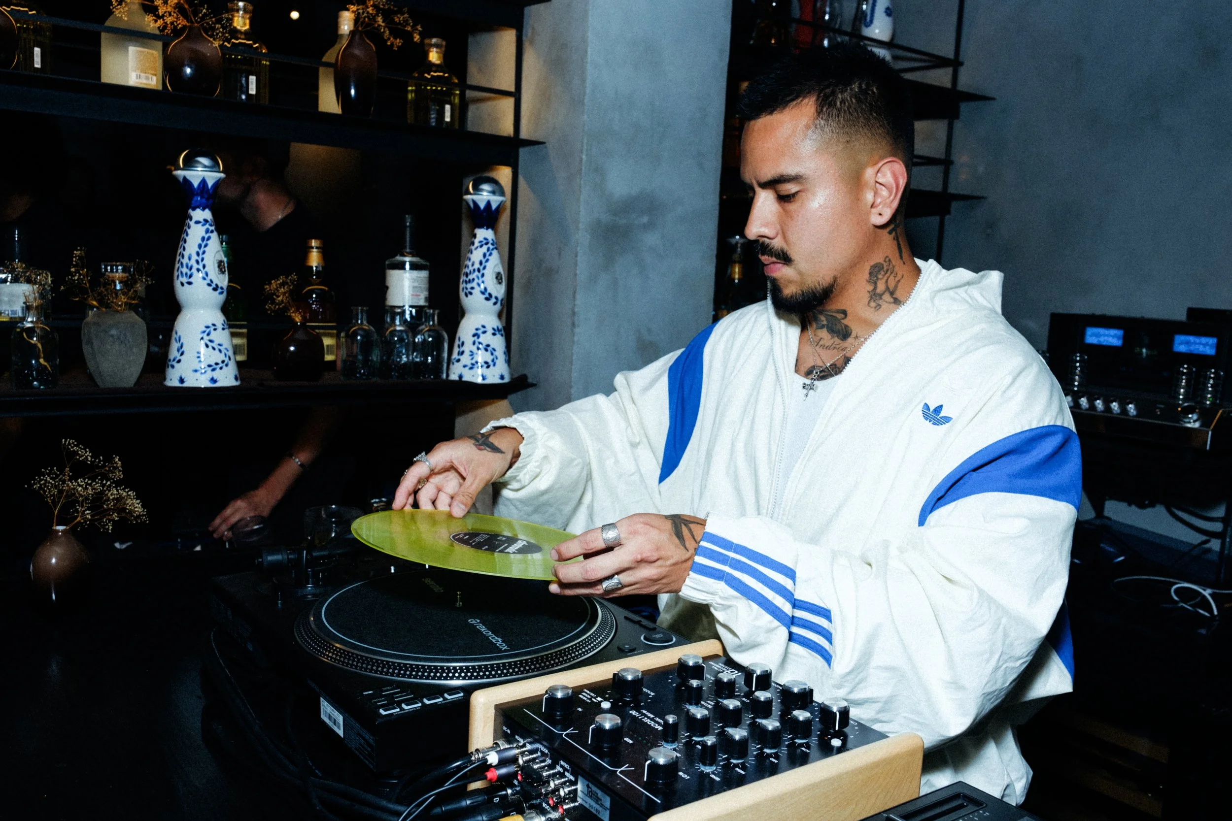 A man with tattoos, short black hair, and a beard, wearing a white Adidas jacket with blue stripes, is DJing with a turntable and mixer in front of him. There are decorative vases and bottles on shelves behind him in a dimly lit room.