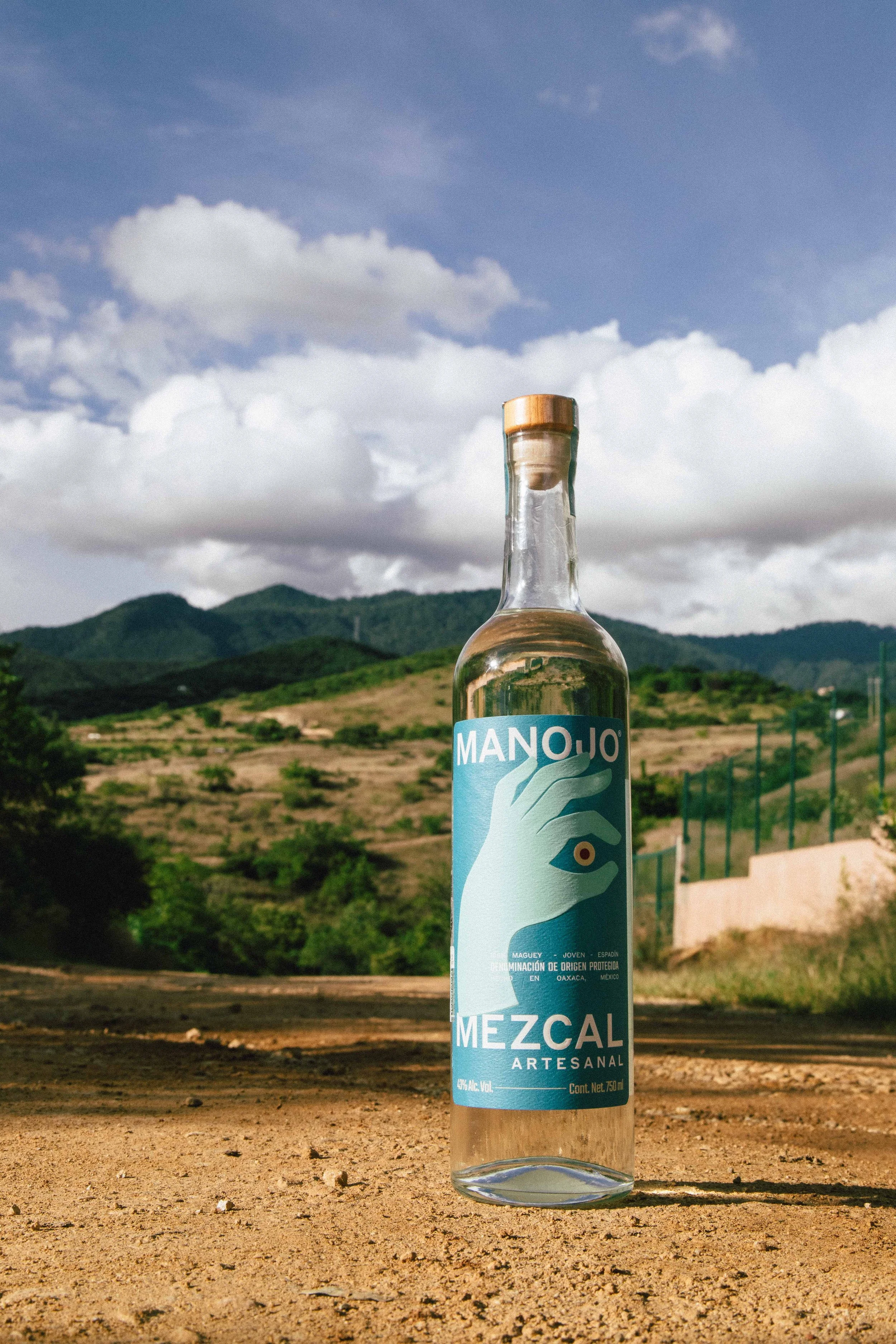 A clear glass bottle of mezcal artisanal spirit with a blue label, placed on dry, cracked soil outdoors with a background of mountains, green hills, and a partly cloudy sky.