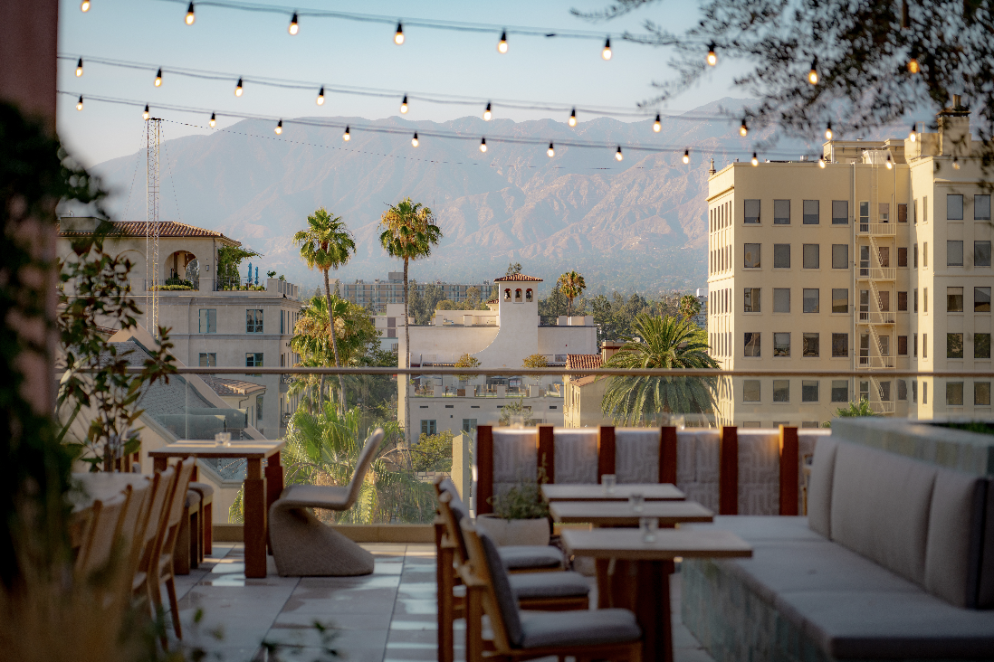 Outdoor terrace with string lights, wooden tables, chairs, and a sofa, overlooking city buildings, palm trees, and mountains in the background during daylight.