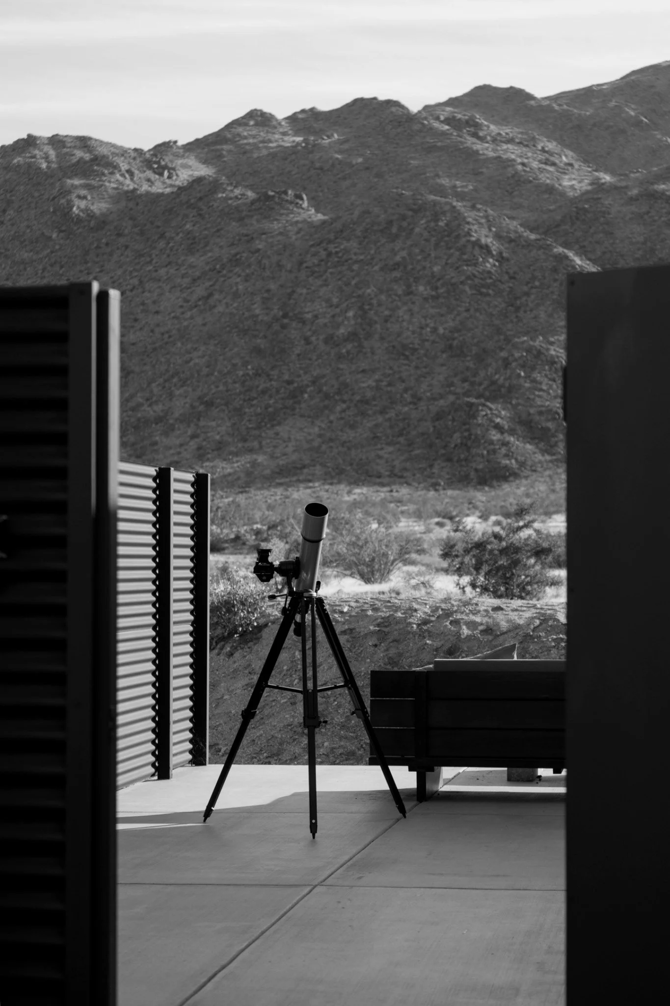 View of a mountain range with a telescope set up outdoors and a building structure in the foreground.
