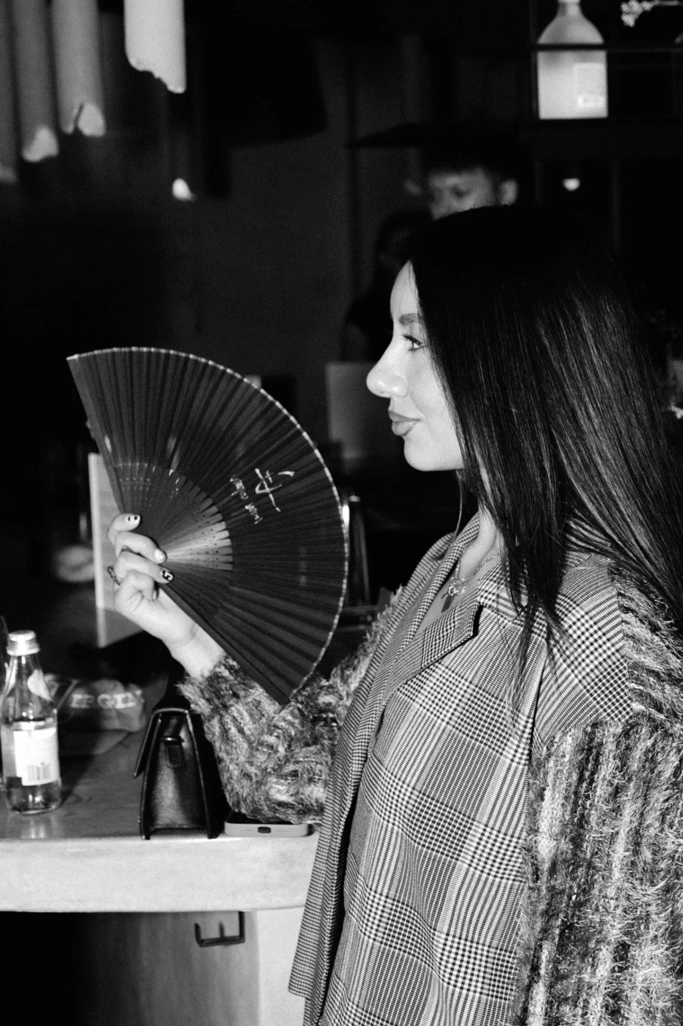 A woman with long dark hair is holding a fan and standing in a room, facing right. She is wearing a checkered blazer over a patterned shirt and is near a table with bottles and a black purse.