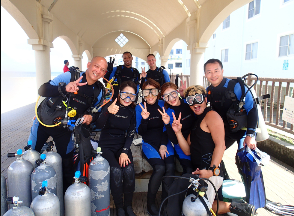 Group of people in scuba diving gear under a covered walkway, smiling and making peace signs.