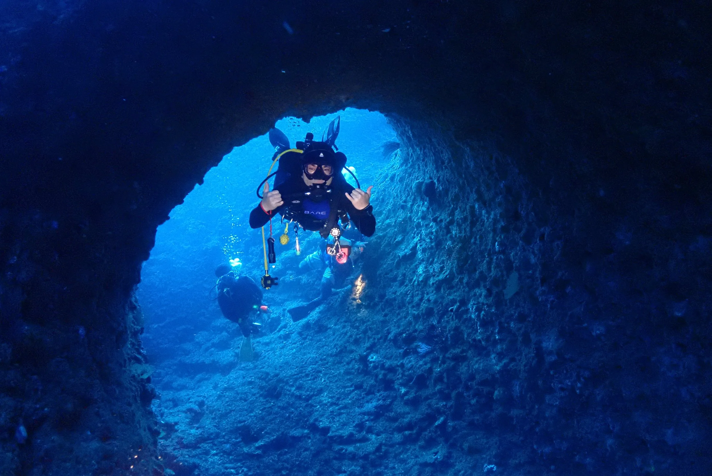 A scuba diver in a cave with blue water, wearing a wetsuit, mask, and gear, making a hand gesture while exploring underwater.