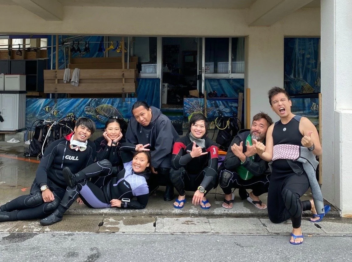 A group of seven people in wetsuits posing happily outside a building, with equipment bags and diving gear in the background.