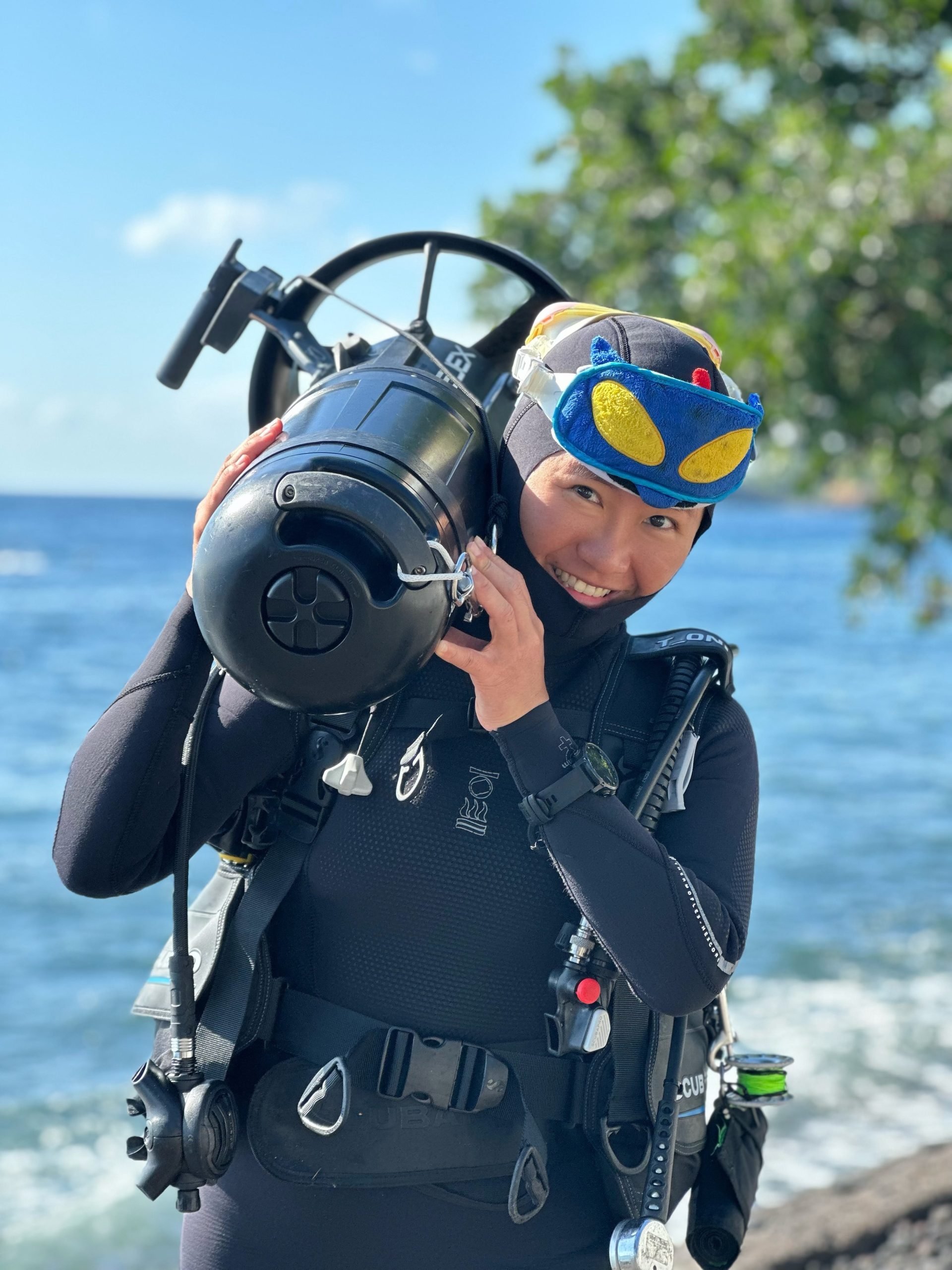 A smiling woman in a wetsuit holding scuba diving gear including a regulator, mask, and tank on a beach near the ocean with trees and a blue sky in the background.