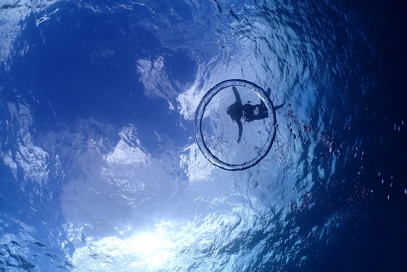 A person snorkeling underwater, viewed from below with sunlight shining through the water, and a circular bubble ring around the snorkeler.