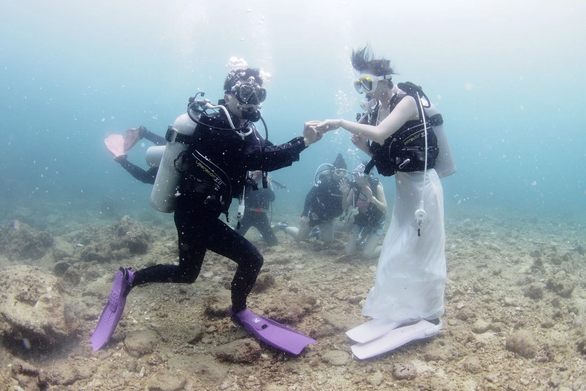 Two women in scuba diving gear underwater, holding hands and interacting; one woman is kneeling on the rocky seabed, the other standing with white fins, both wearing masks and breathing apparatus, with a group of divers in the background.