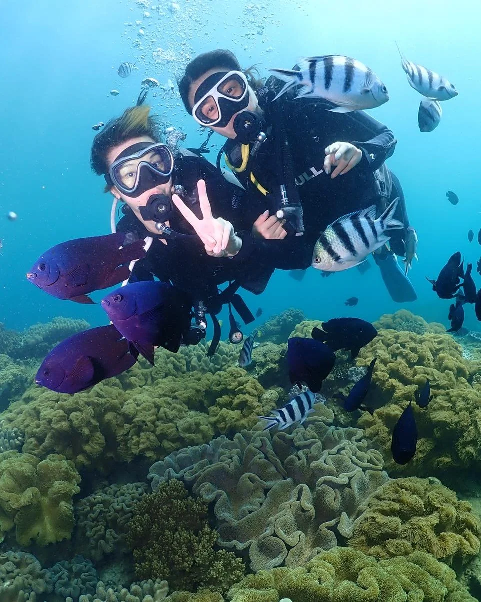 Two scuba divers underwater surrounded by colorful fish and coral reefs, making peace signs.
